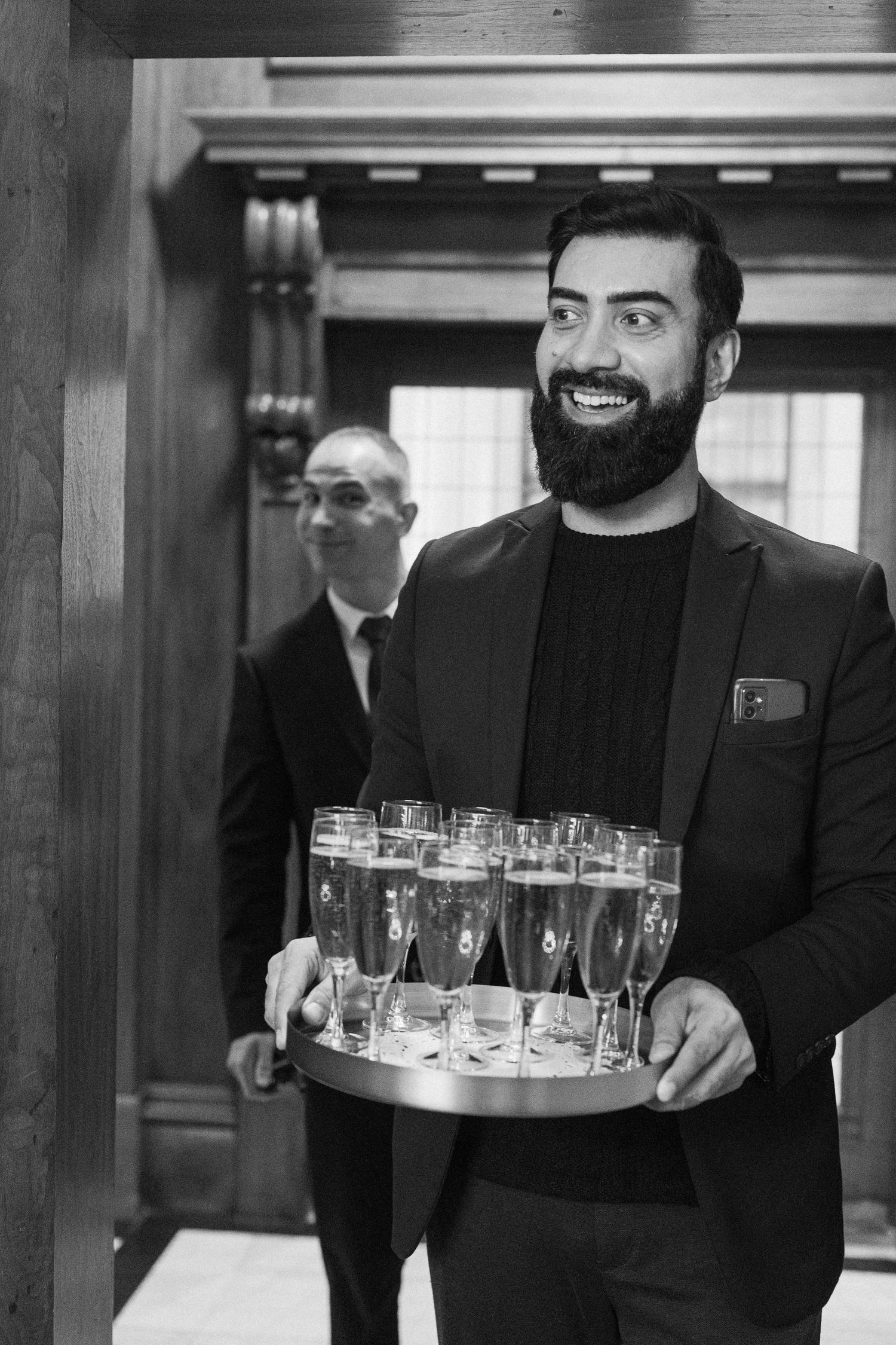 Smiling bearded waiter carrying tray of champagne glasses at wedding reception in black and white.