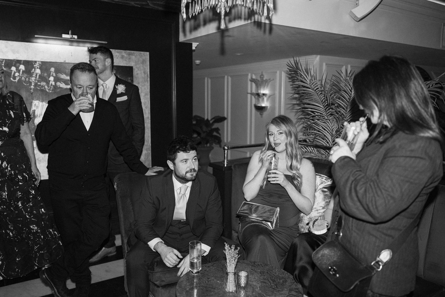 Black and white photo of wedding guests socializing and laughing together in a cozy bar setting.