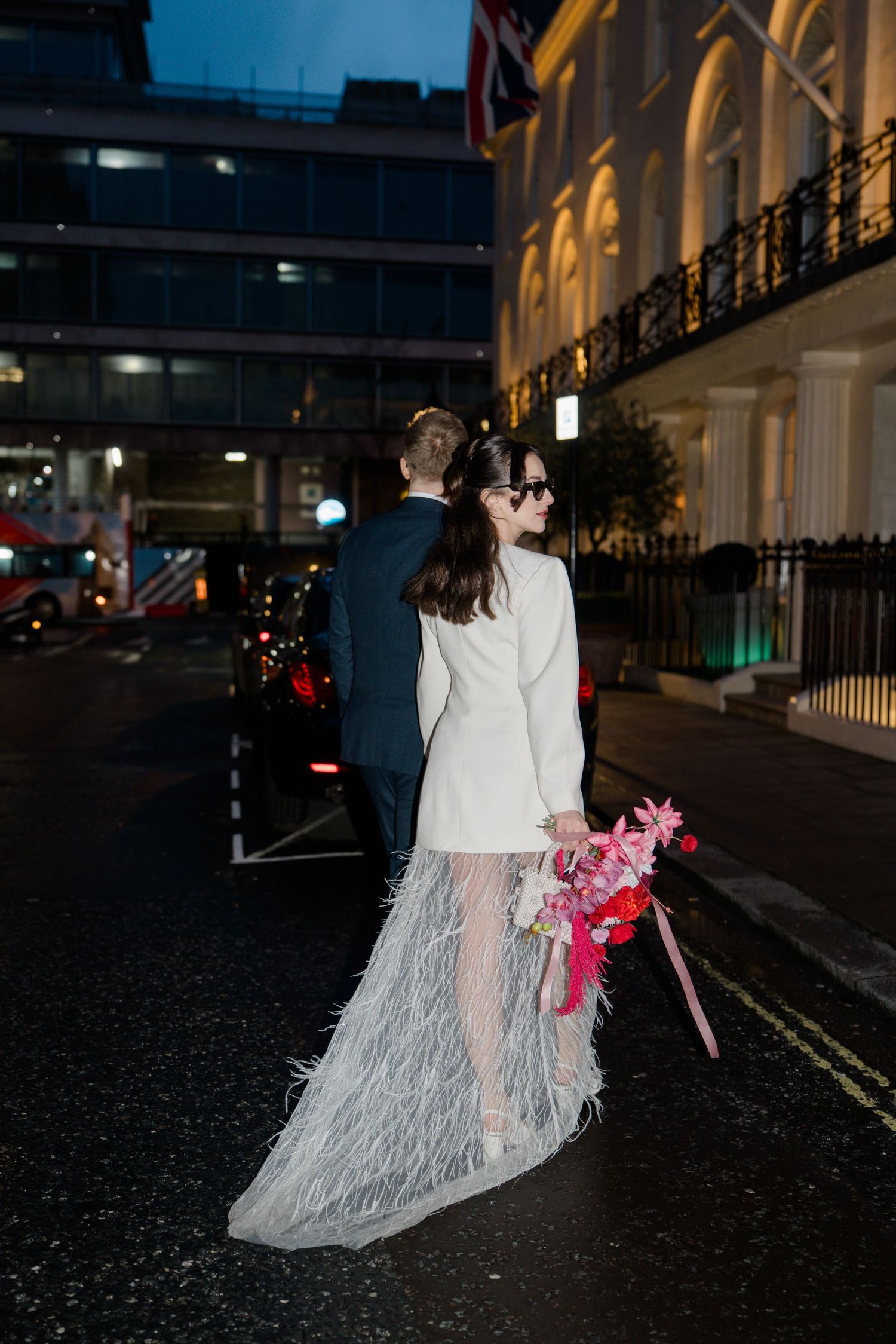 Bride in white blazer and feather skirt with groom on London street at night.