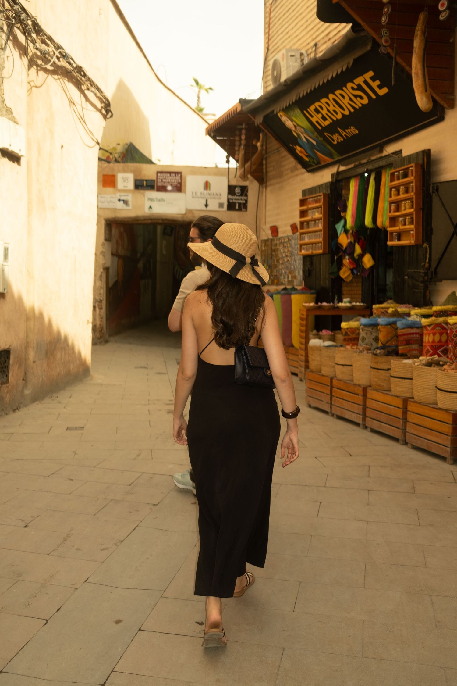 Woman in black dress and straw hat walking through a Moroccan medina market alley past a herborist shop.