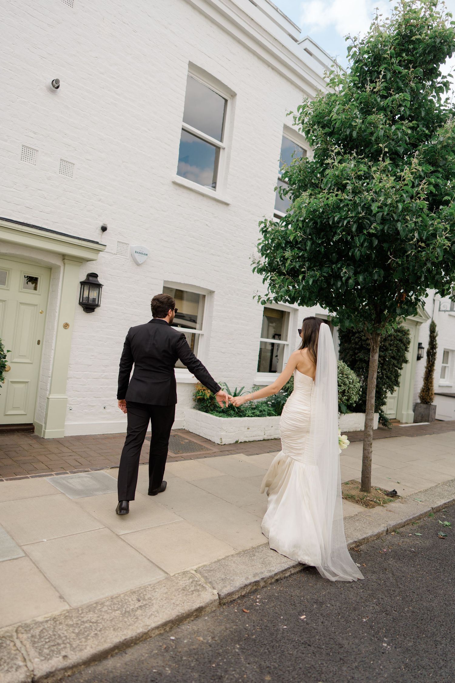 Bride and groom holding hands walking along a sidewalk outside a white brick building on their wedding day.