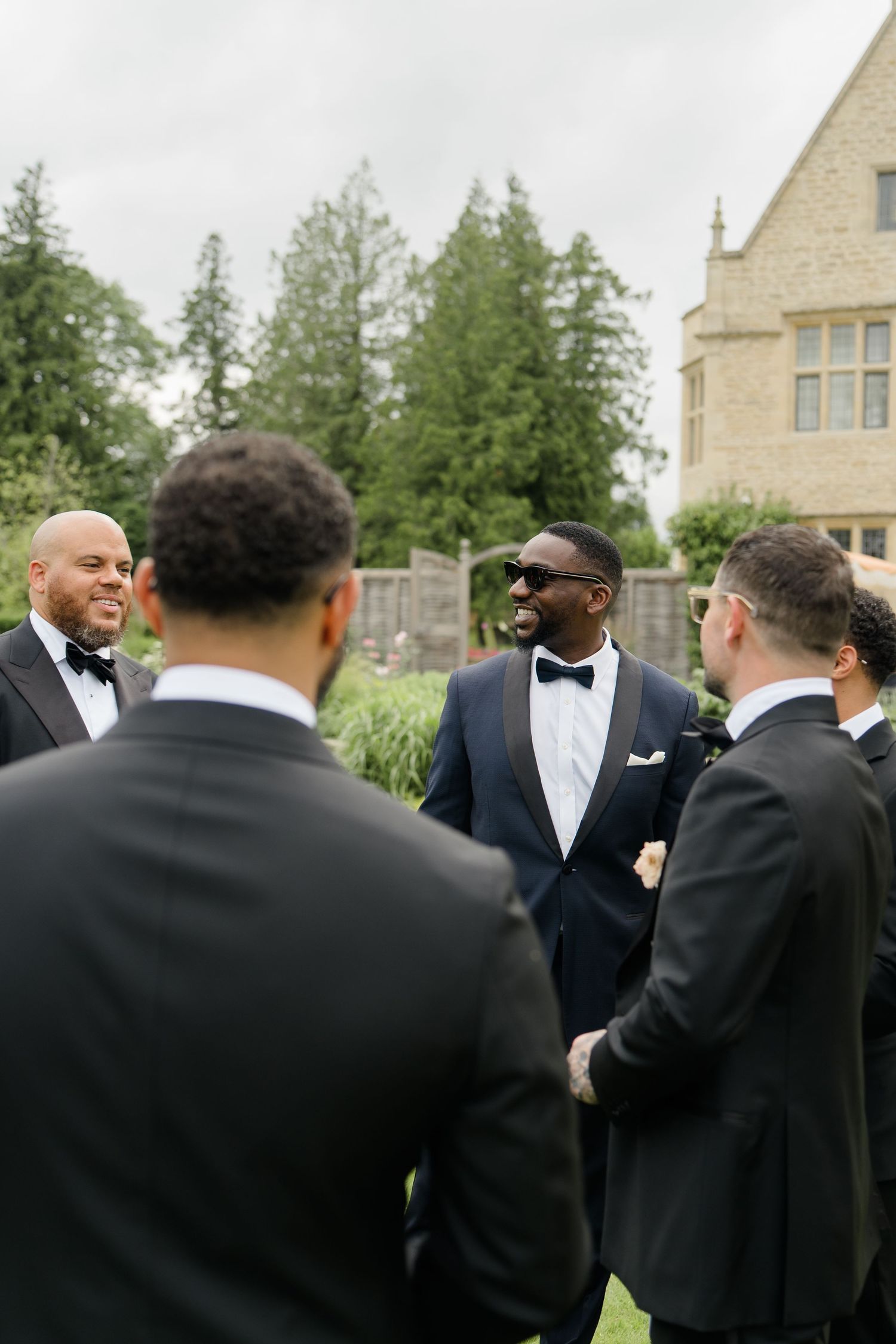 Groom in navy tuxedo with sunglasses smiling with groomsmen in black suits outdoors.