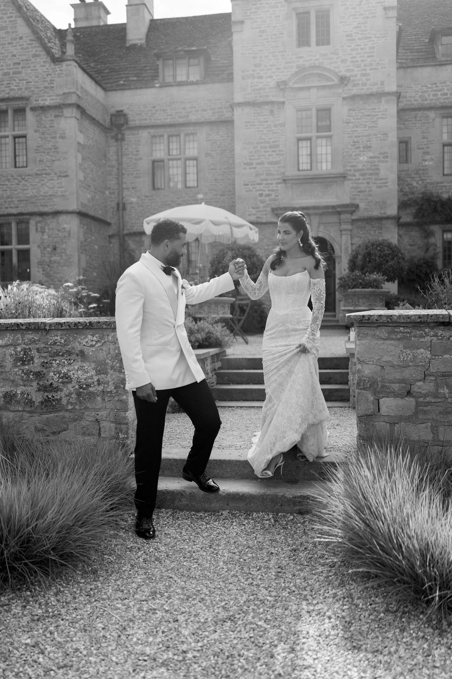 Black and white photo of bride and groom dancing under umbrella outside a grand manor house.