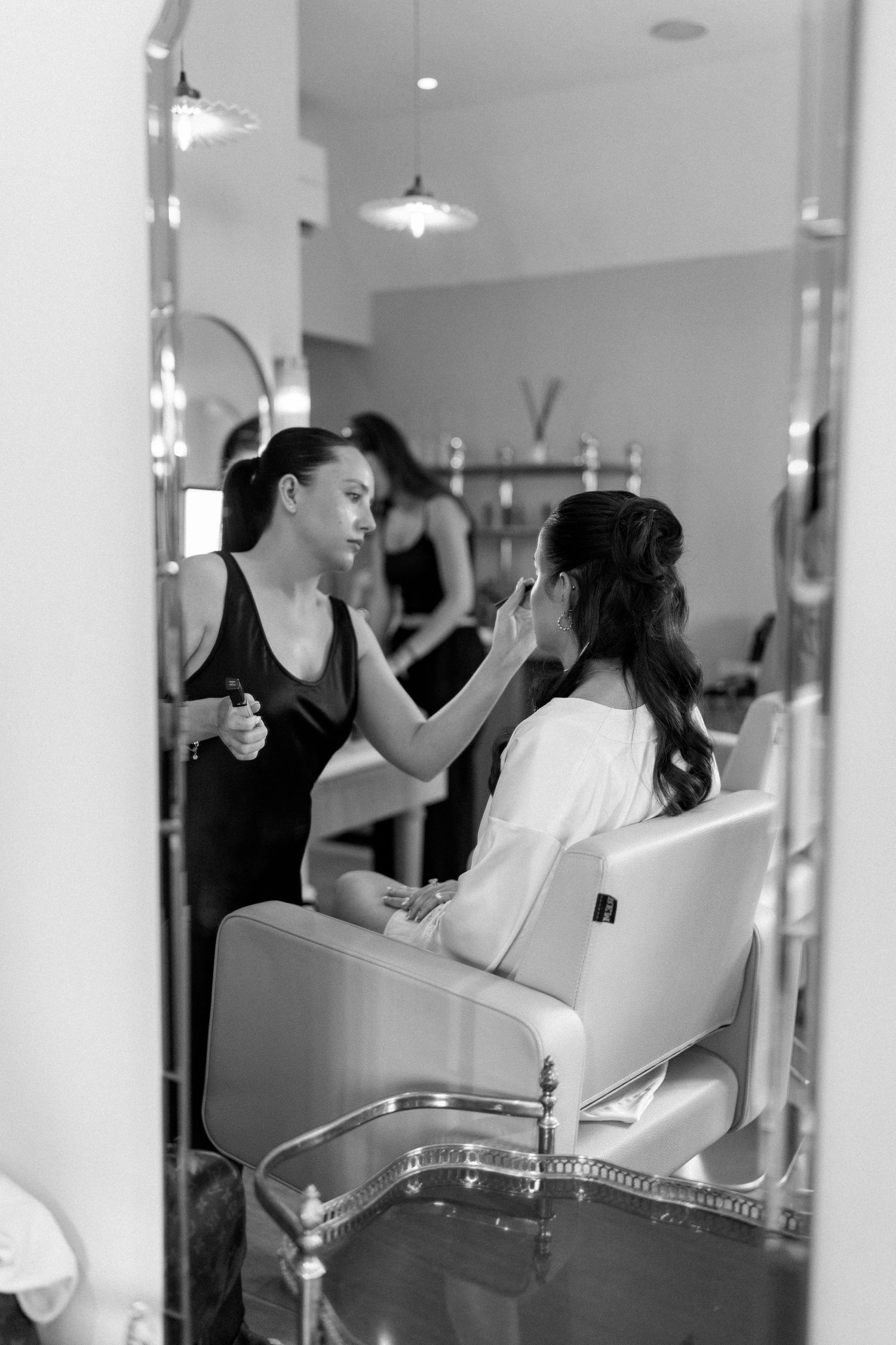 Makeup artist applying makeup to a bride in a salon mirror reflection, black and white wedding preparation photo.