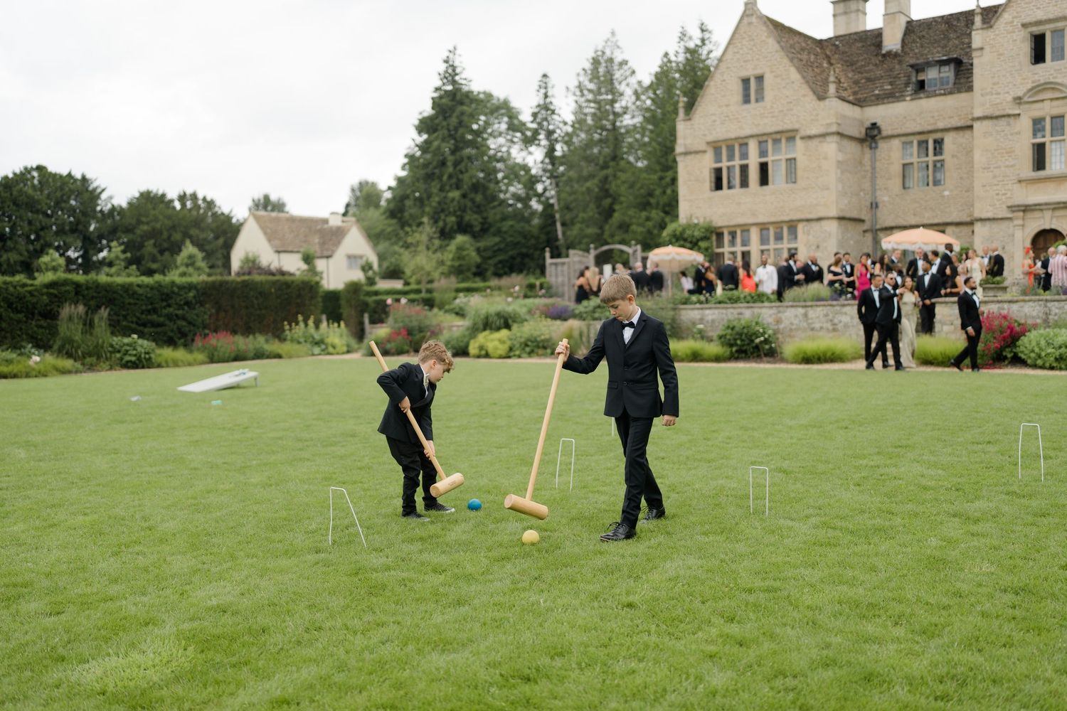 Two boys in tuxedos playing croquet on lawn outside a grand stone manor wedding venue.