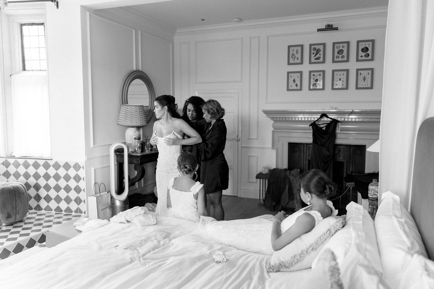 Bride being helped into wedding dress by bridesmaids in elegant room with fireplace, black and white.