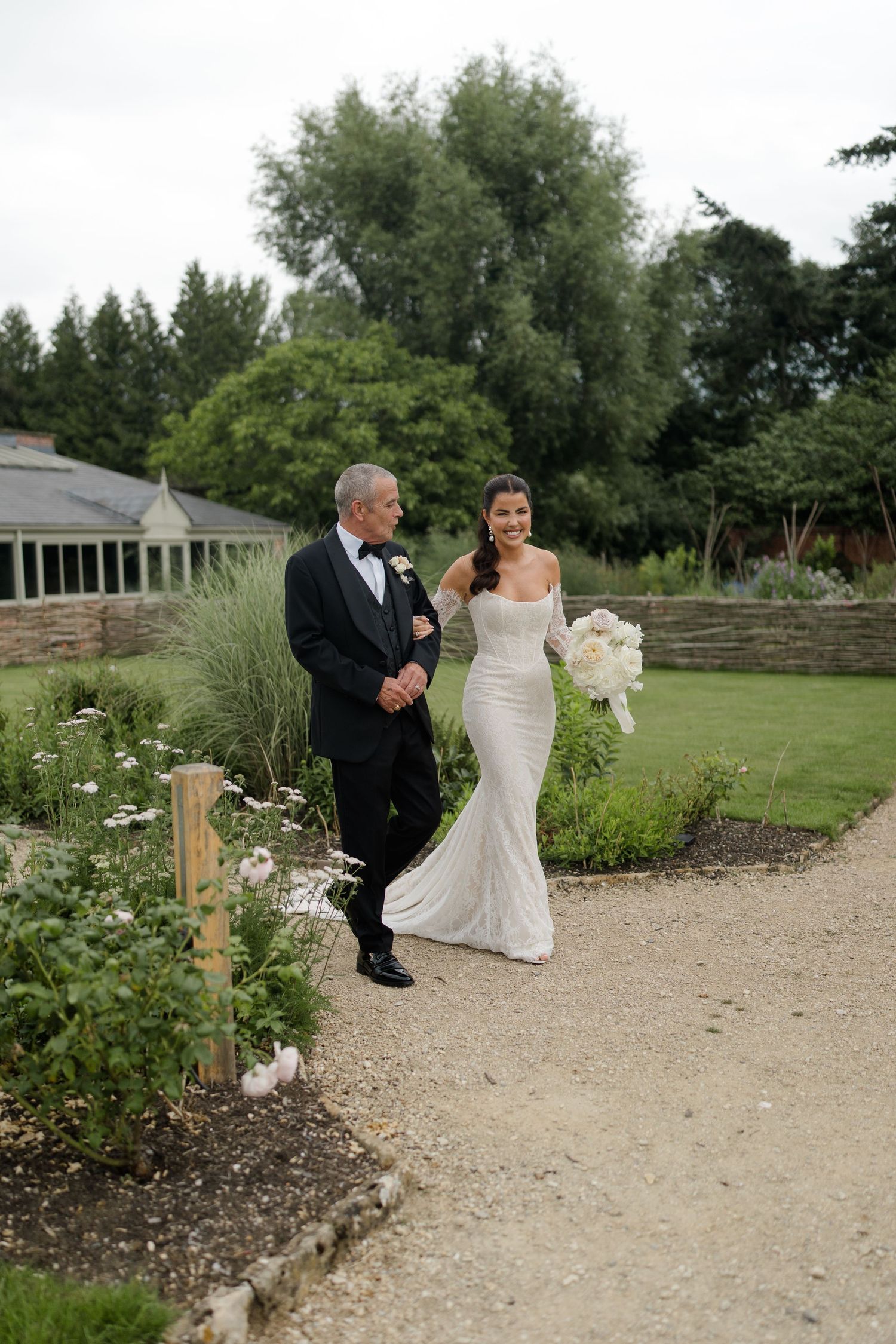 A bride in a fitted white lace gown walks with an older gentleman in a tuxedo along a garden path.