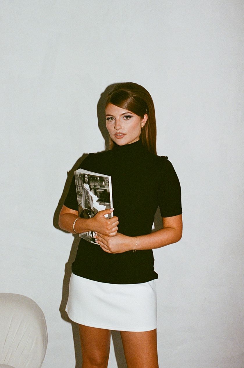 Young woman in black turtleneck top and white mini skirt holding a book, posing against a white wall.