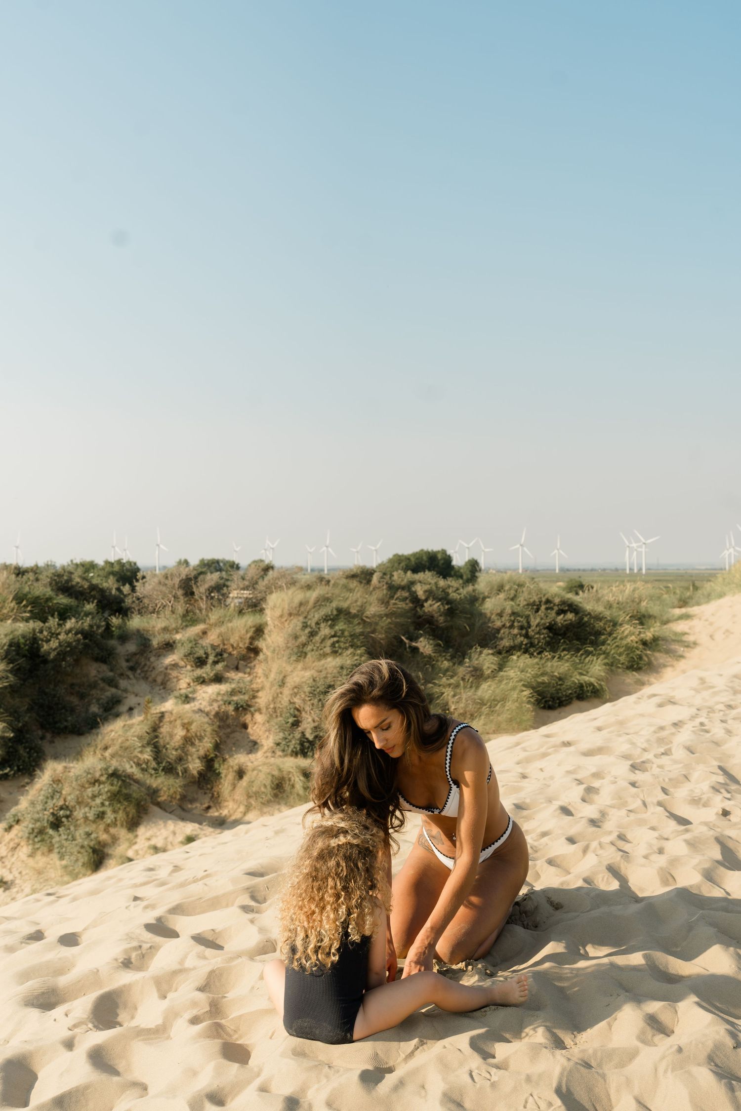 Two people sitting together on sandy beach dunes with vegetation in background.