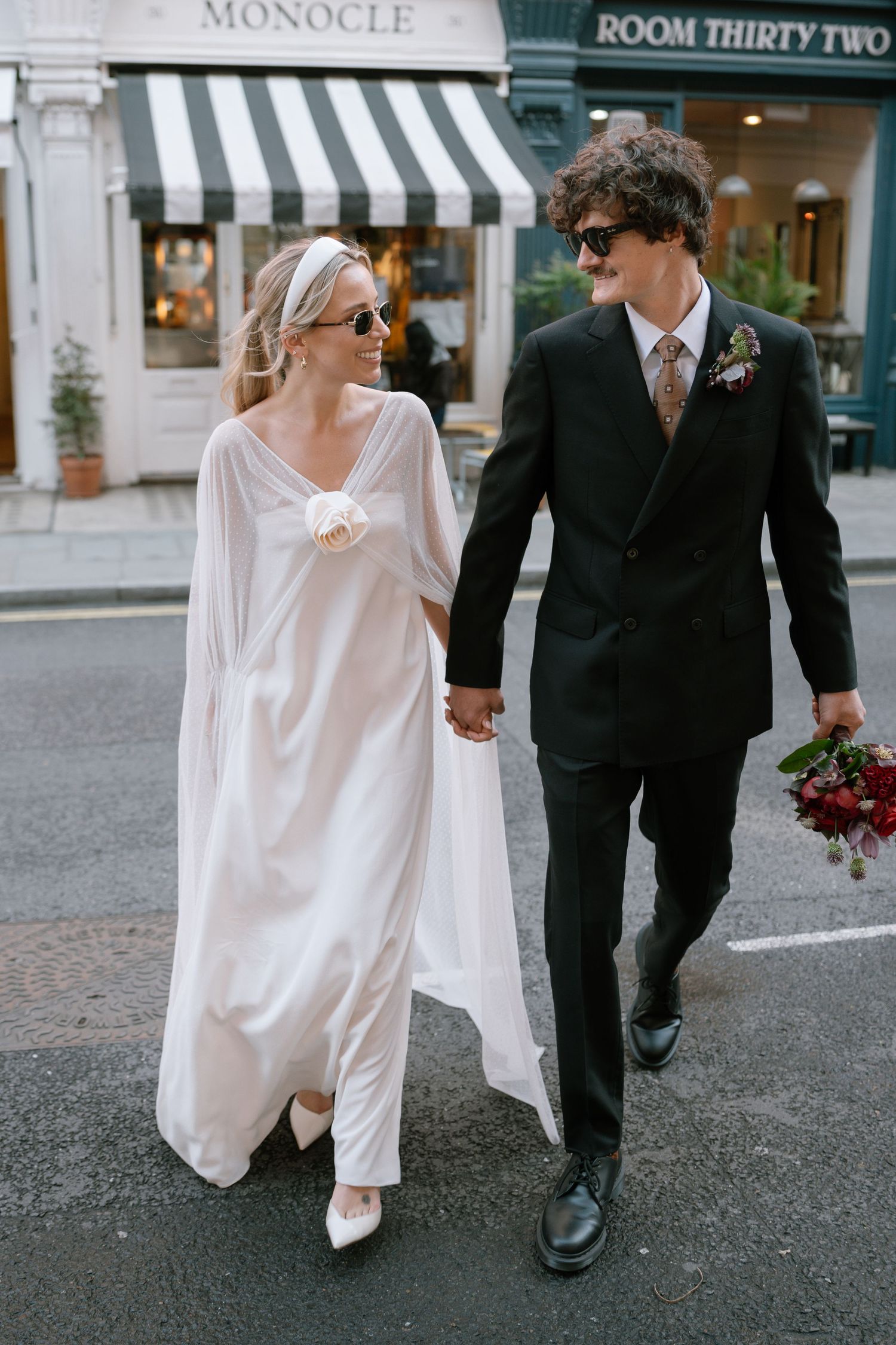 Newlyweds stroll down a cobblestone street past boutique shops in an urban setting.