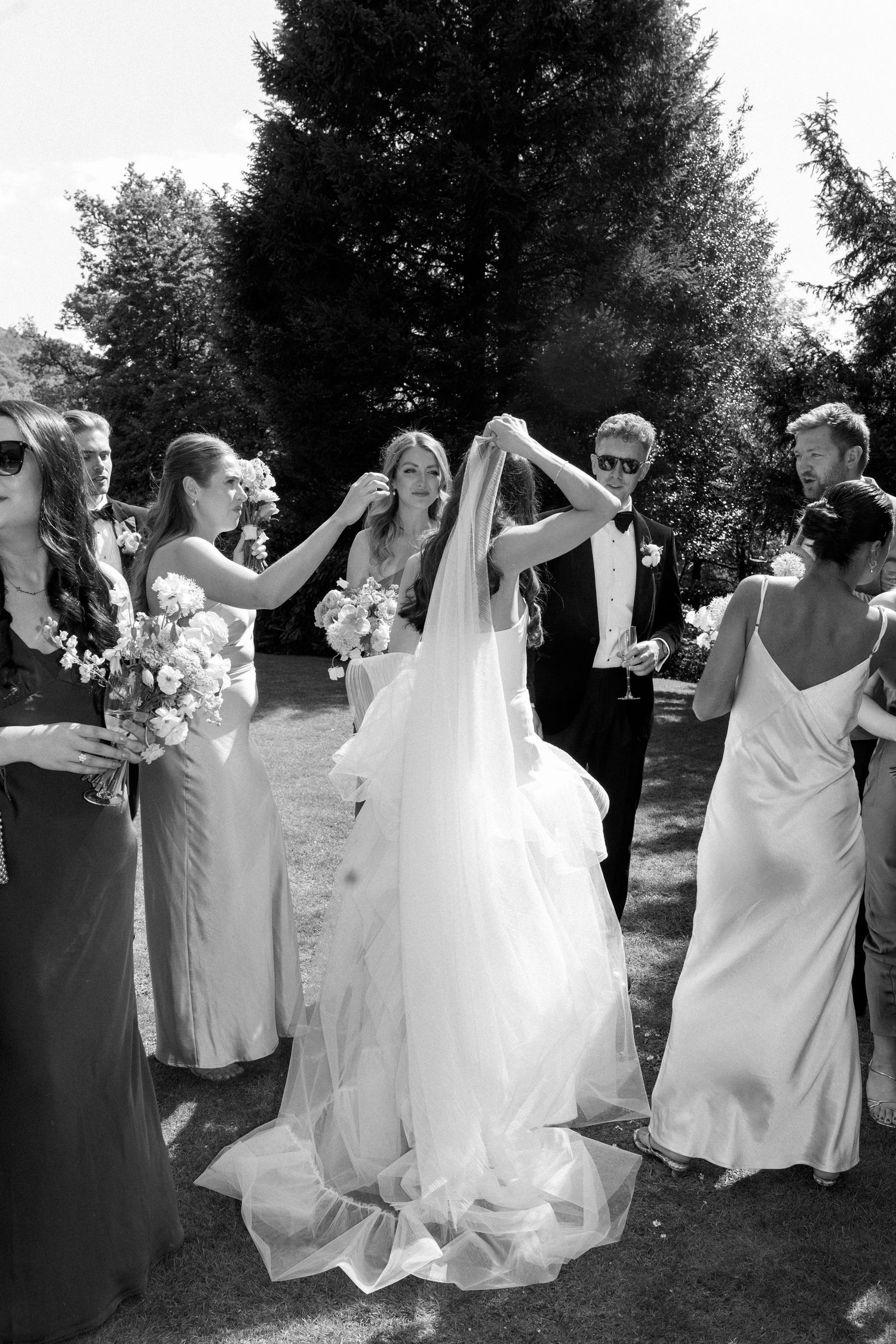 Black and white photo captures bridesmaids helping with bride's veil during outdoor celebration.