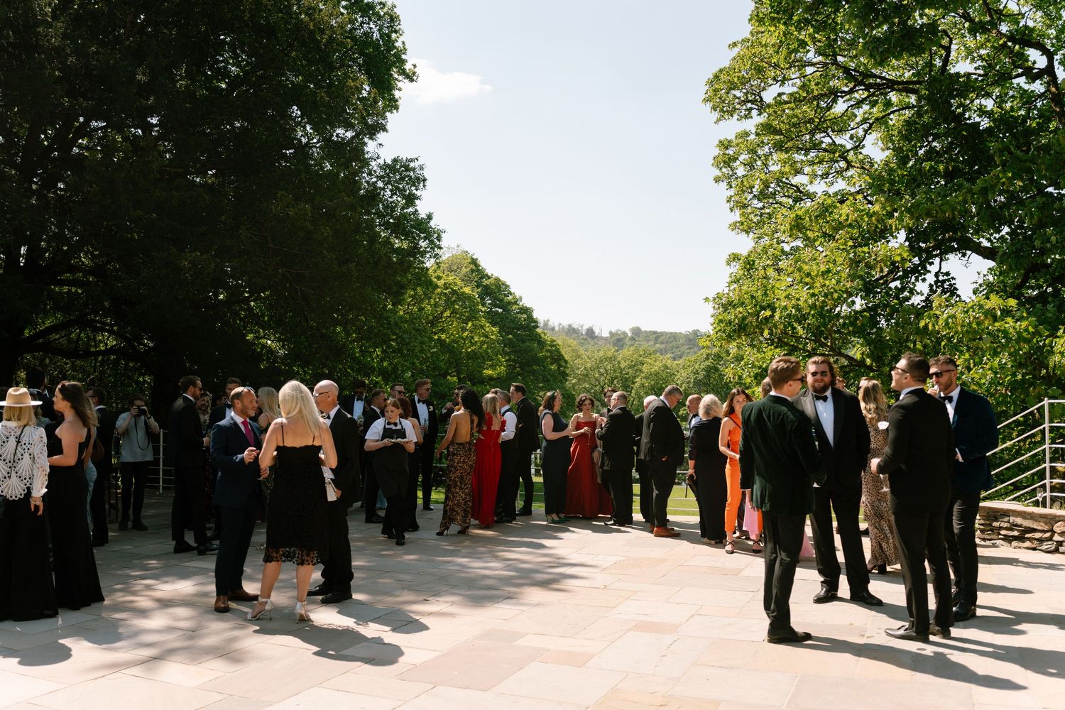 Guests socialize at an outdoor wedding reception on a sunny day surrounded by green trees.