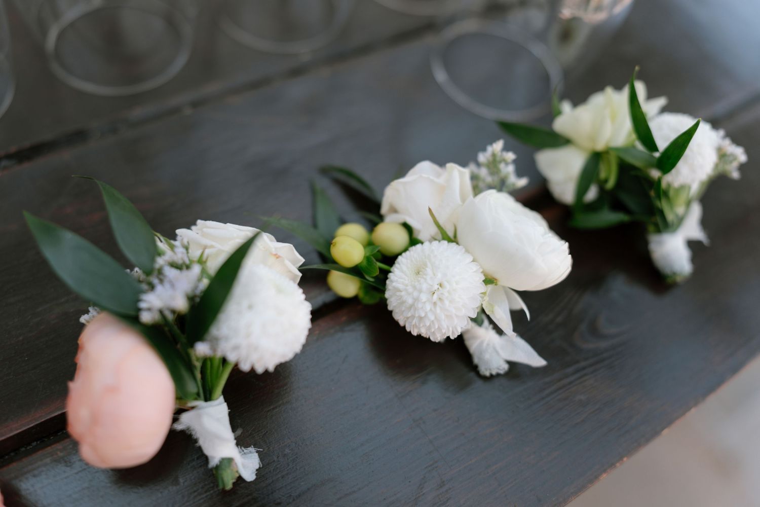 White floral boutonnieres with greenery arranged on dark surface.