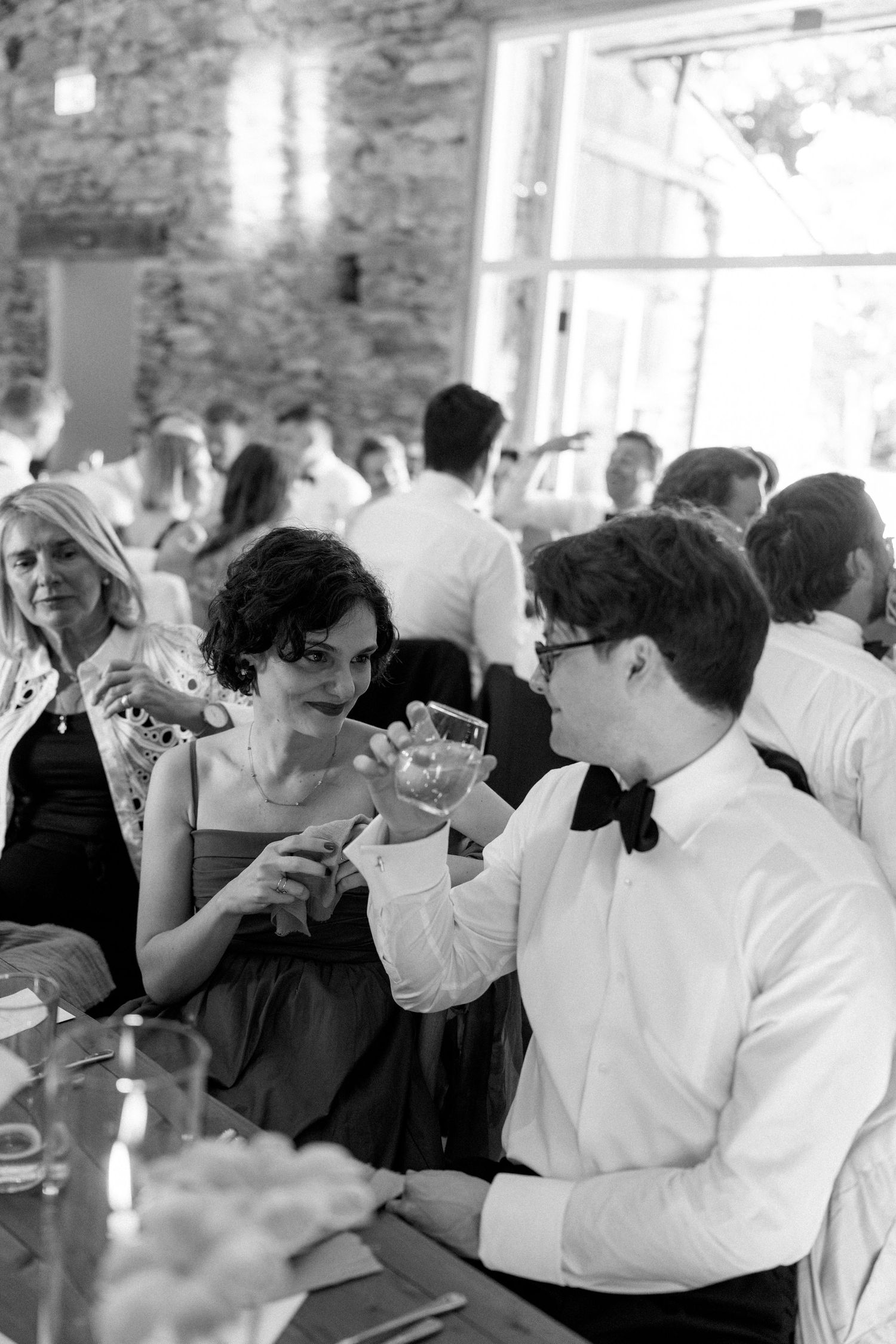 Black and white photo of wedding guests mingling at an elegant reception with stone wall backdrop.