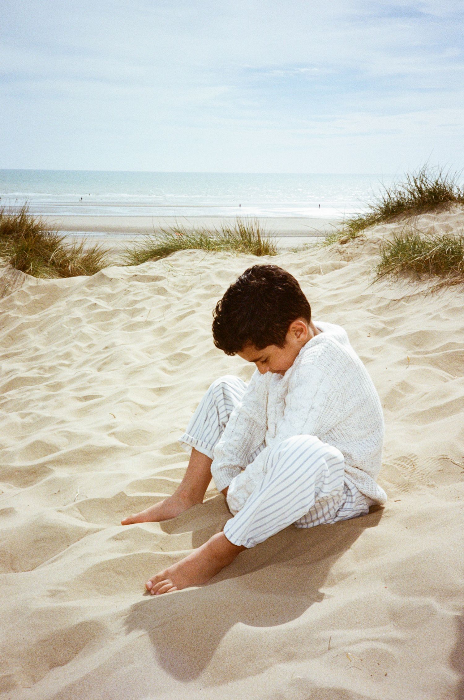 A young boy in white striped clothing sits on sandy beach dunes, playing with sand, with the ocean in the background.