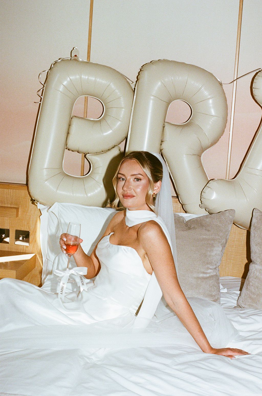 Bride in white gown and veil sits on bed holding wine glass with large letter balloons spelling BRIDE behind her.