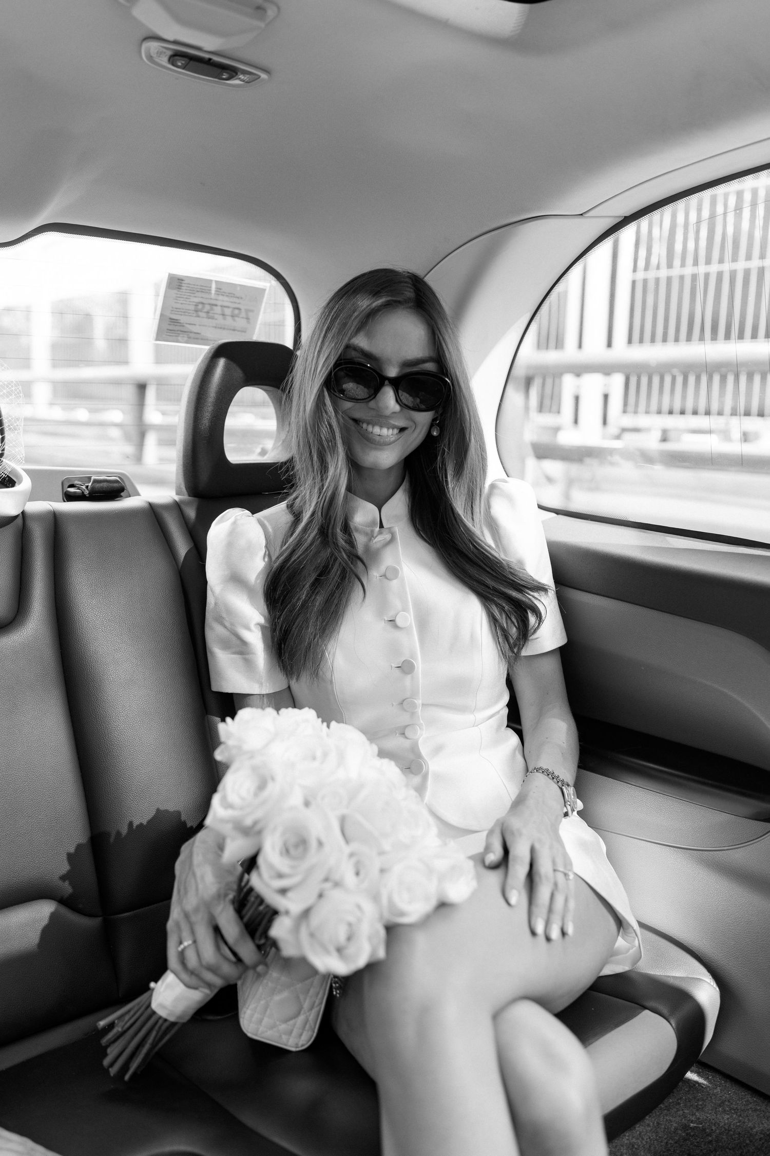 Black and white photo of a smiling bride in white dress and sunglasses holding rose bouquet in car backseat.