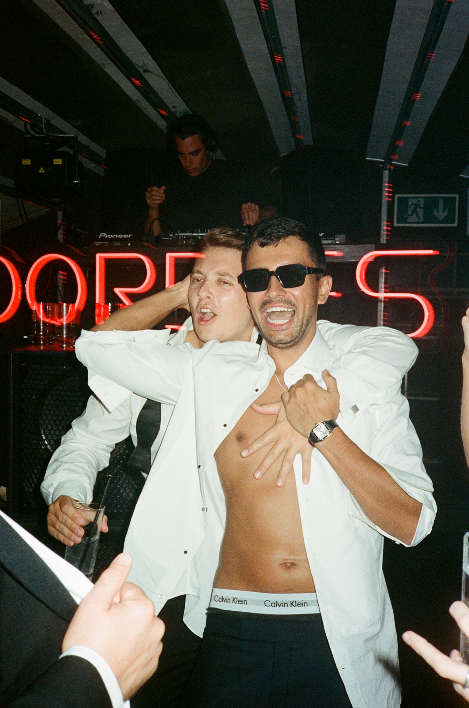 Two men dancing at a nightclub party, one wearing sunglasses with open shirt, DJ performing in background.