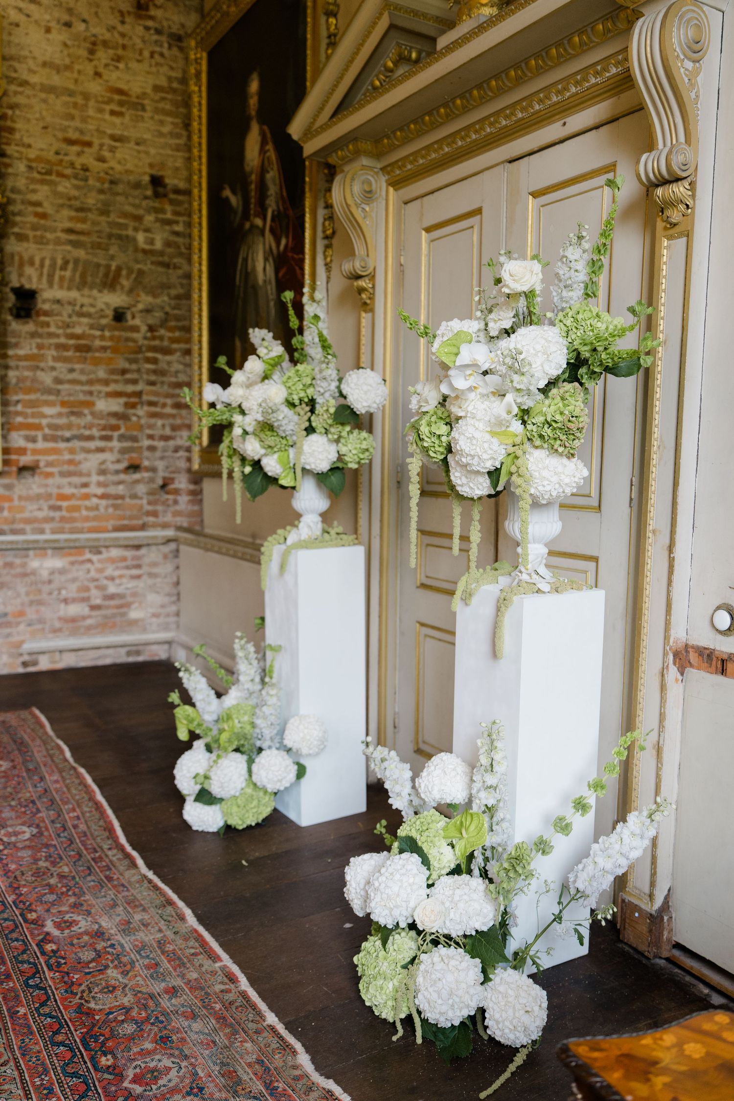 Elegant wedding ceremony entrance with white floral arrangements on pedestals beside ornate golden doors.