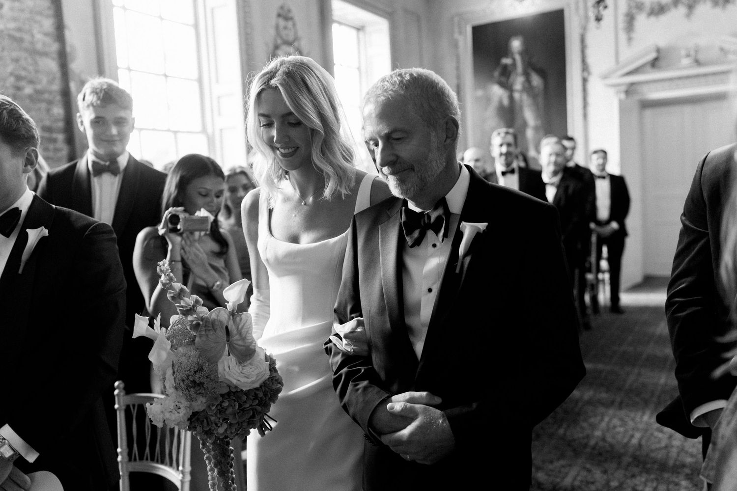 Black and white photo of smiling bride walking down aisle with father at wedding ceremony.