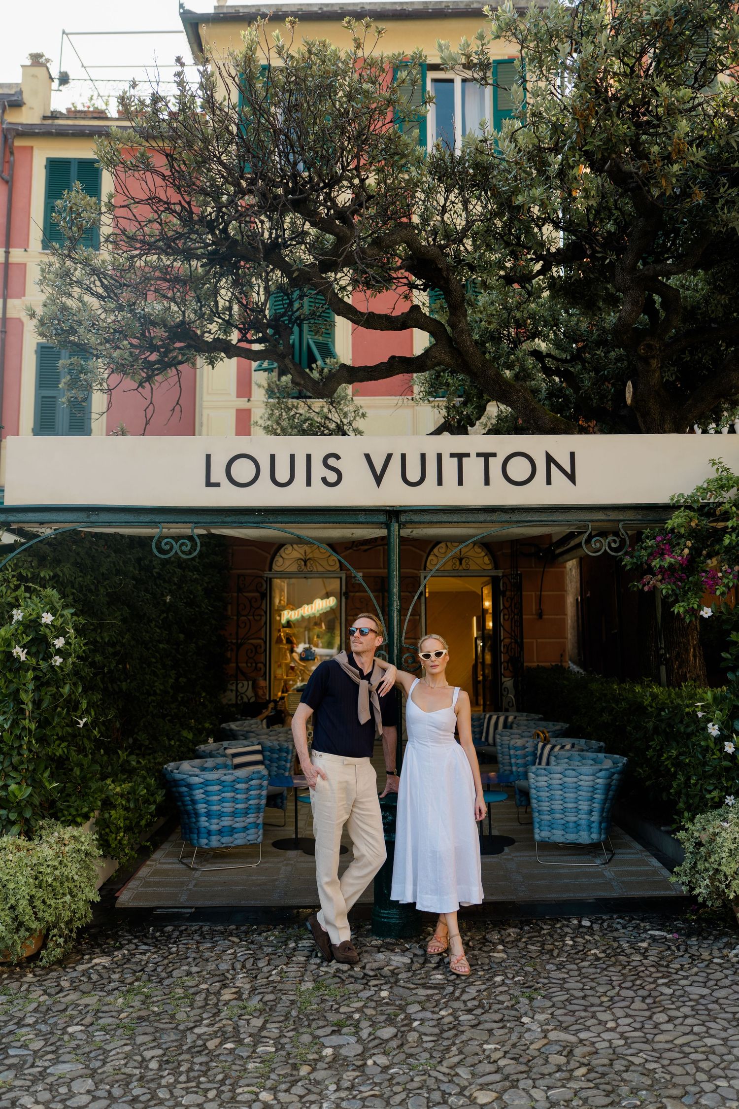 Fashionable couple posing confidently in front of Louis Vuitton boutique surrounded by lush greenery.