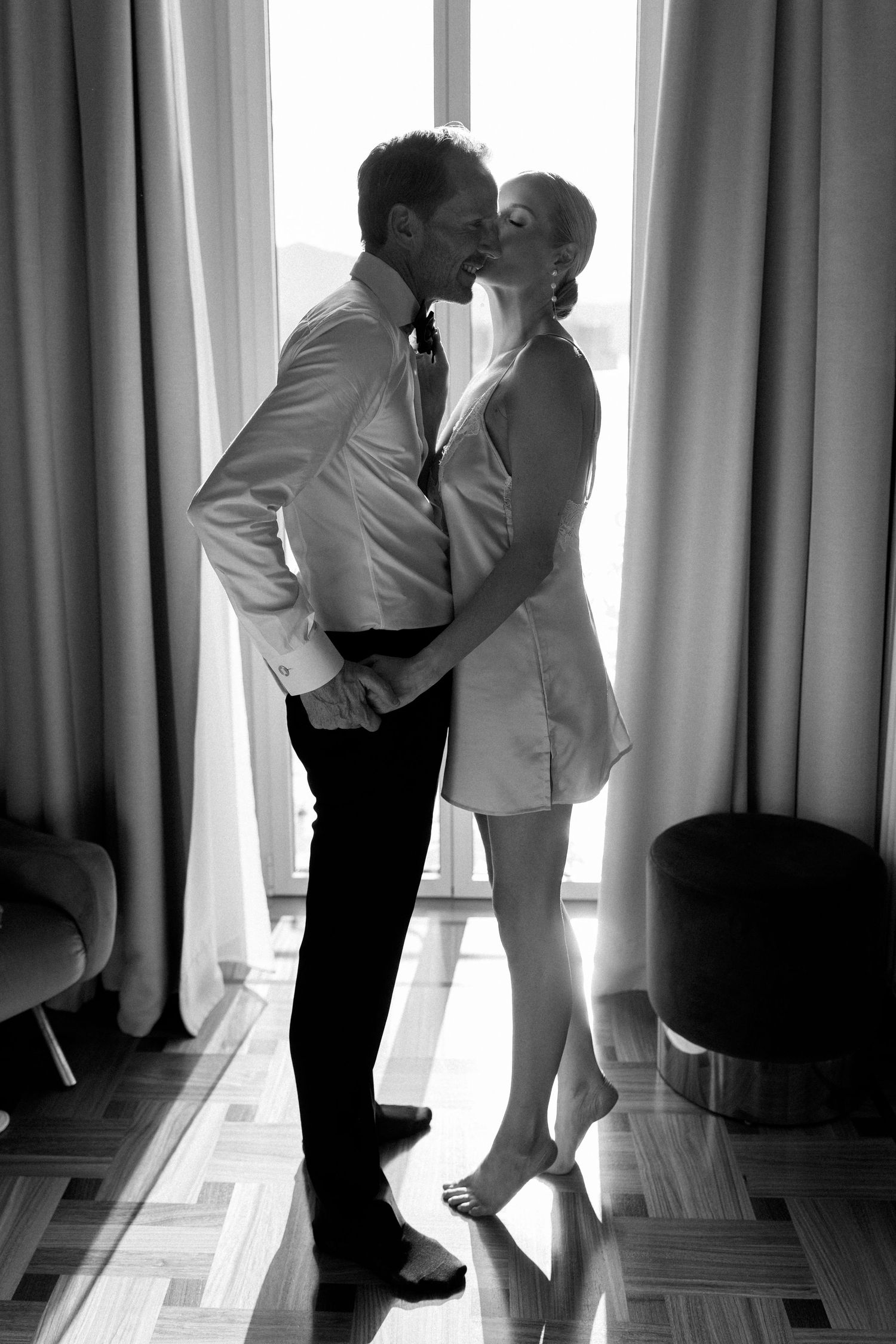 Black and white romantic photo of couple kissing in silhouette by bright hotel room window.