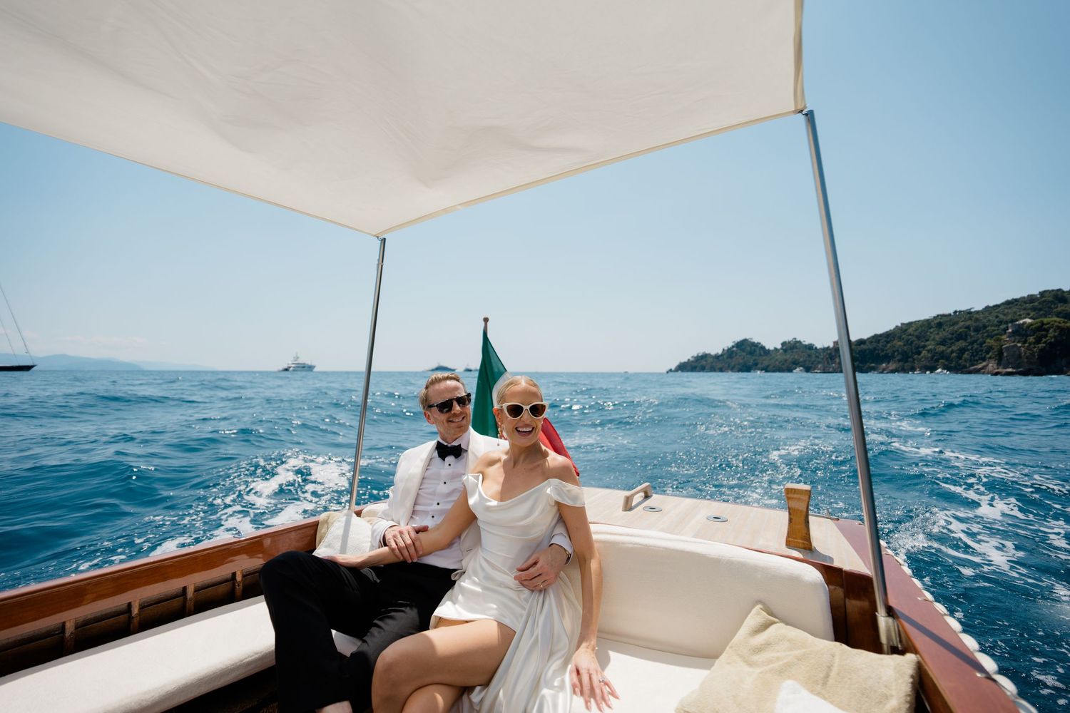 Happy newlywed couple sitting together on luxury wooden boat sailing blue Mediterranean Sea waters.