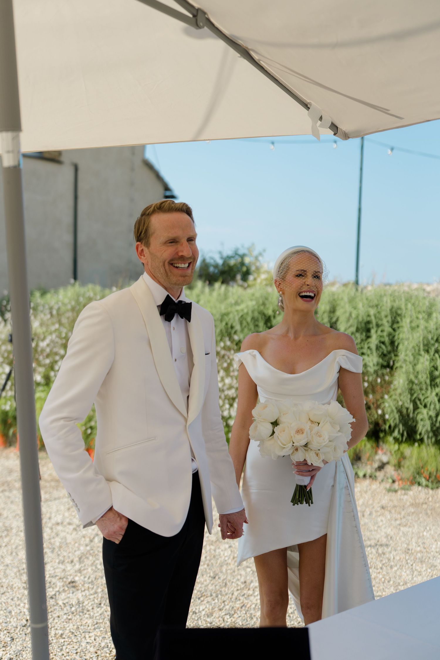 Joyful bride and groom in white tuxedo laughing together at outdoor wedding ceremony.