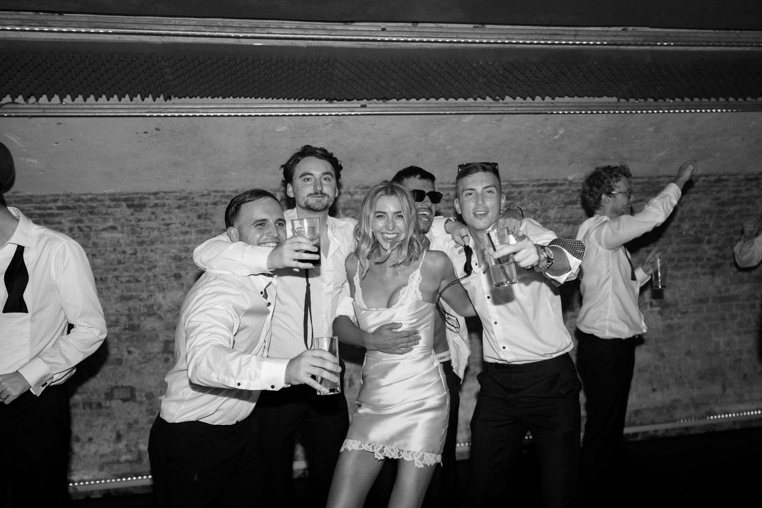 Black and white photo of wedding guests celebrating on dance floor, holding drinks and smiling at a reception party.