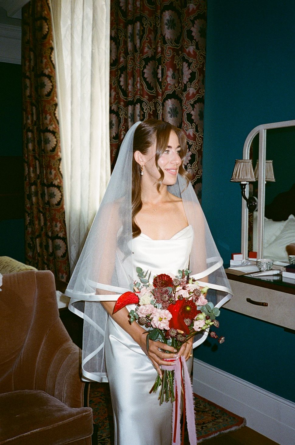 Bride in white satin gown and veil holds red and pink bouquet in elegant room with teal walls.
