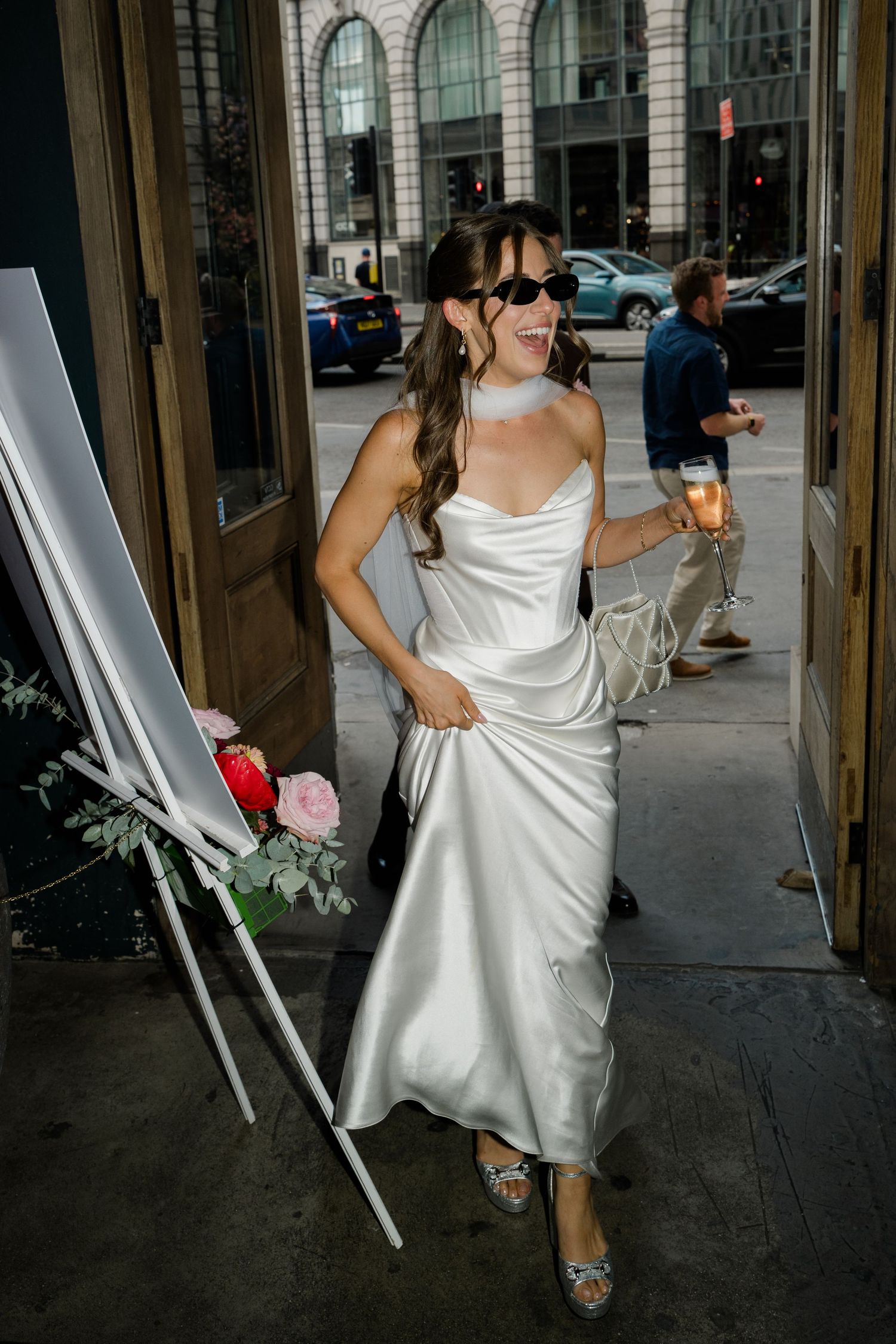 Smiling bride in white satin gown and sunglasses arrives at wedding venue entrance.
