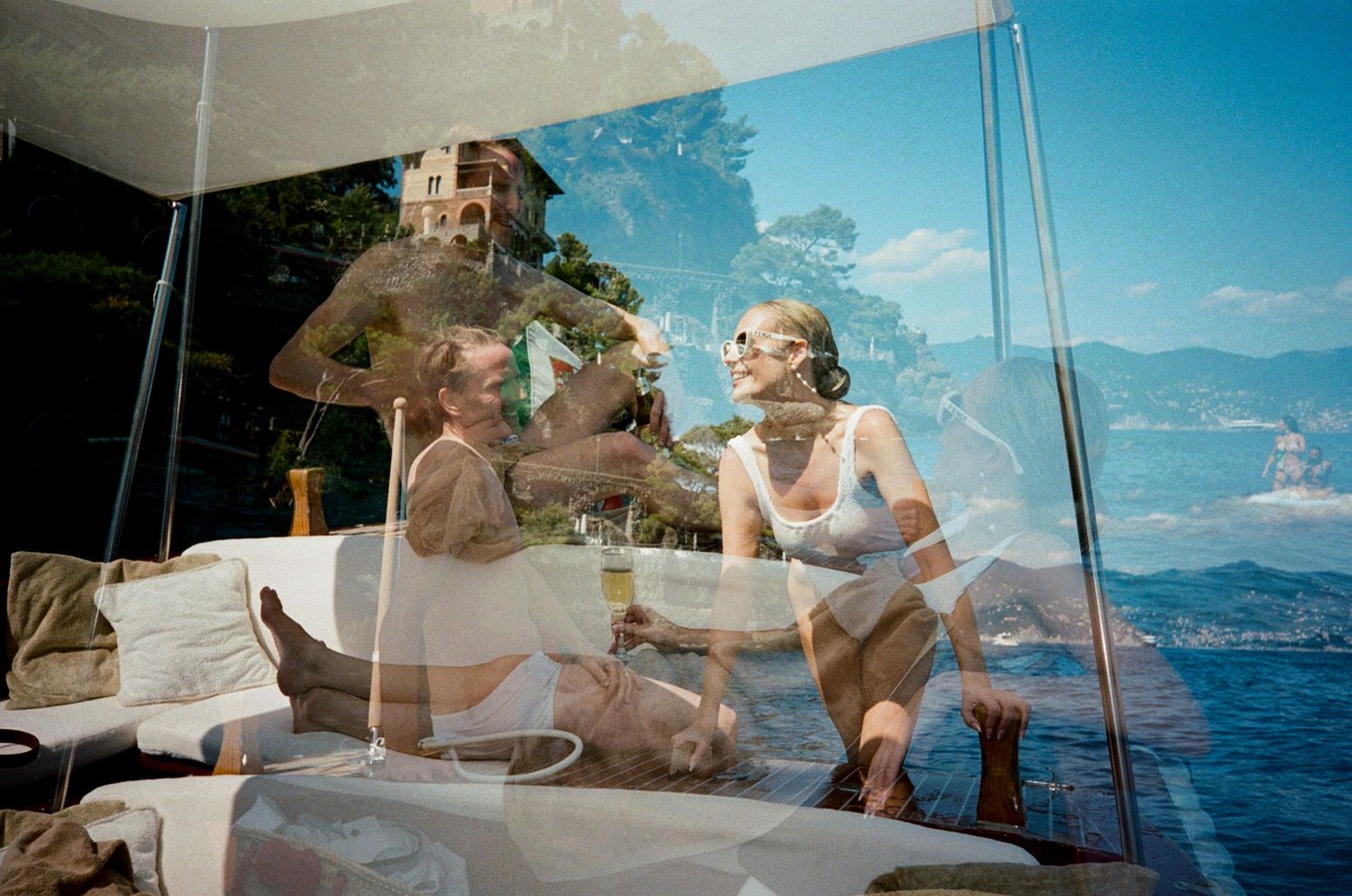 Couple reflected in boat window glass enjoying champagne with scenic Italian lakeside villa and mountains in background.