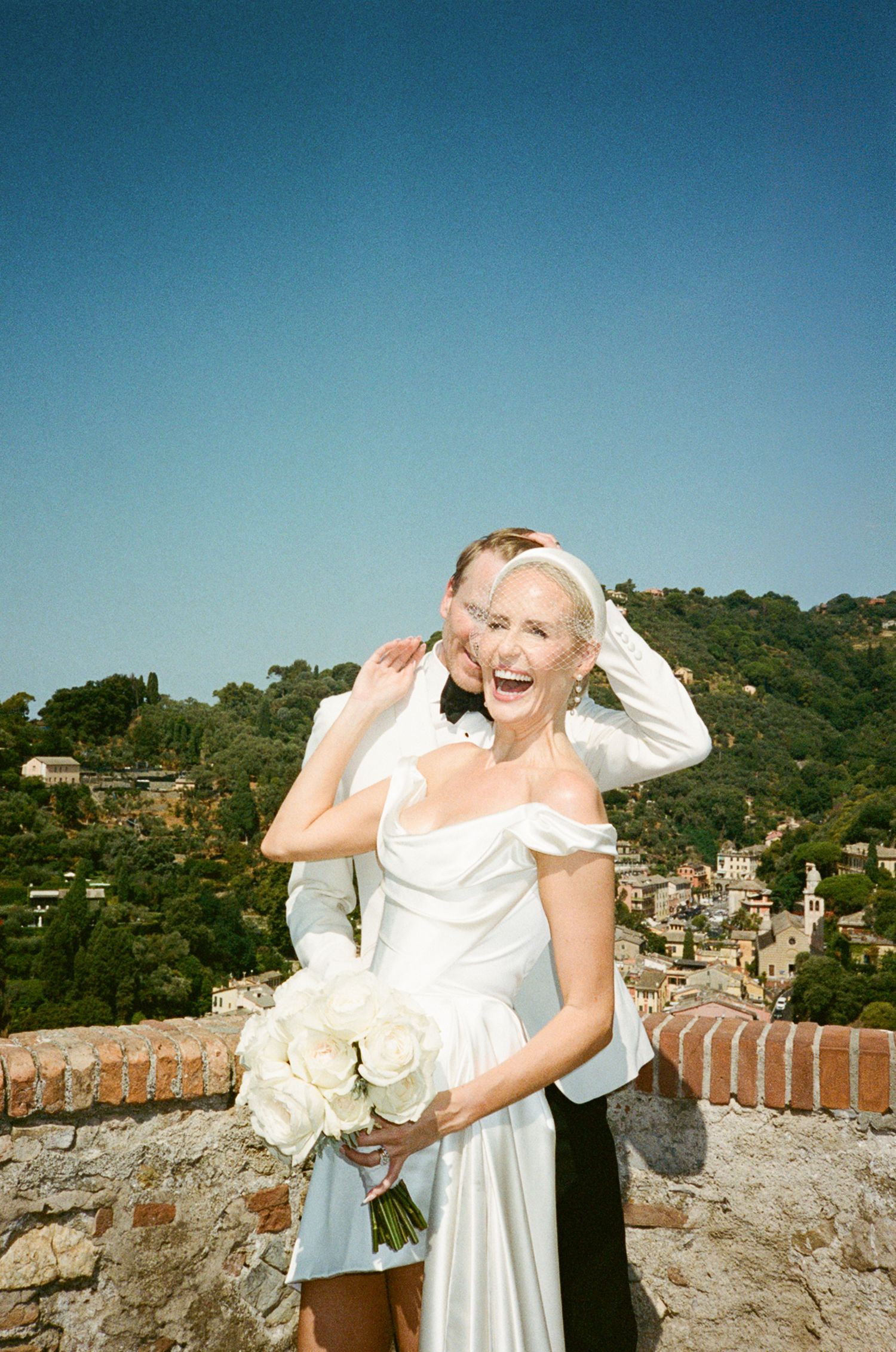 Joyful bride in white off-shoulder gown holds rose bouquet with groom behind her, scenic hillside village backdrop.