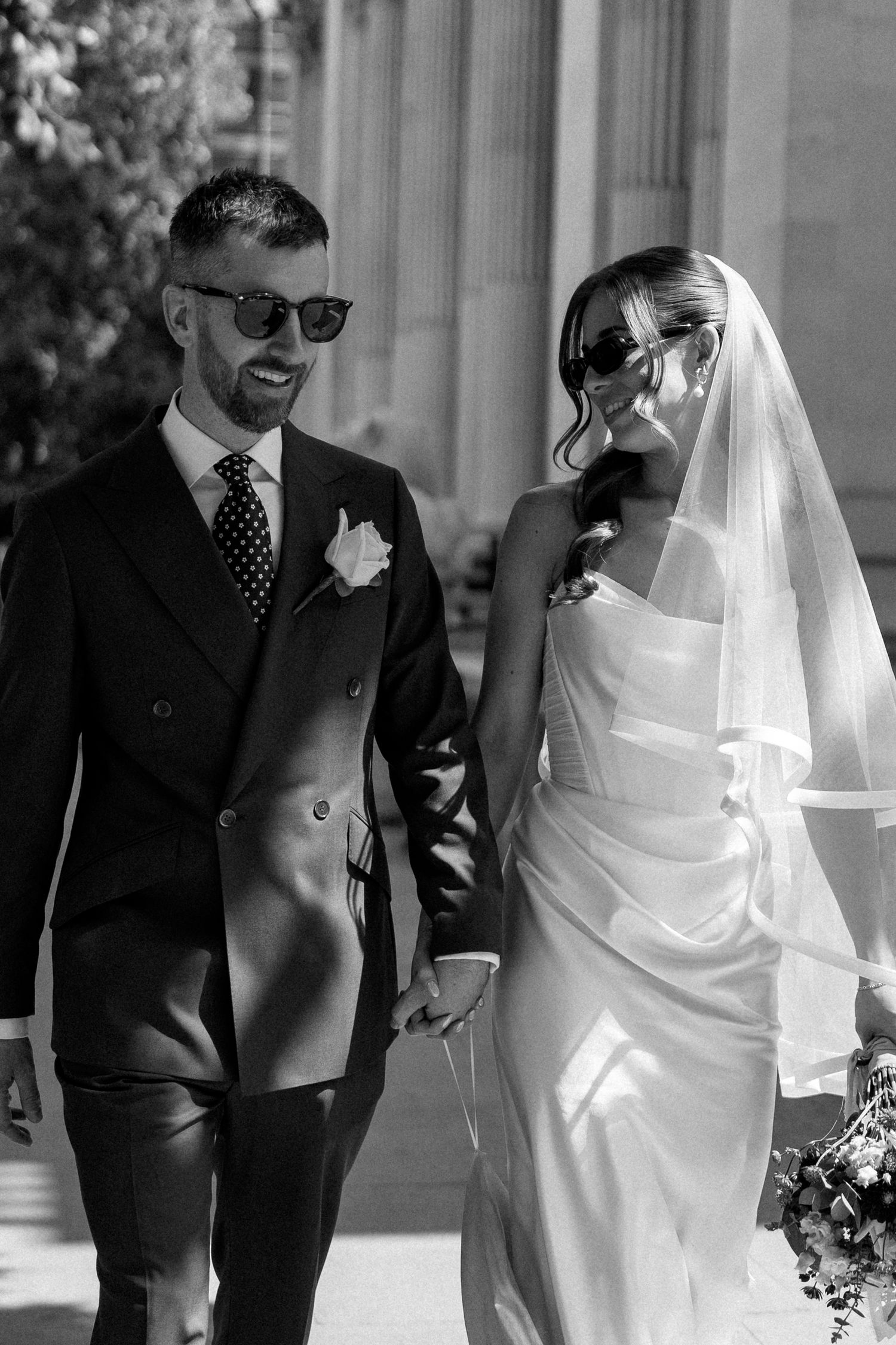 Stylish bride and groom wearing sunglasses walking hand in hand outside in black and white photo.