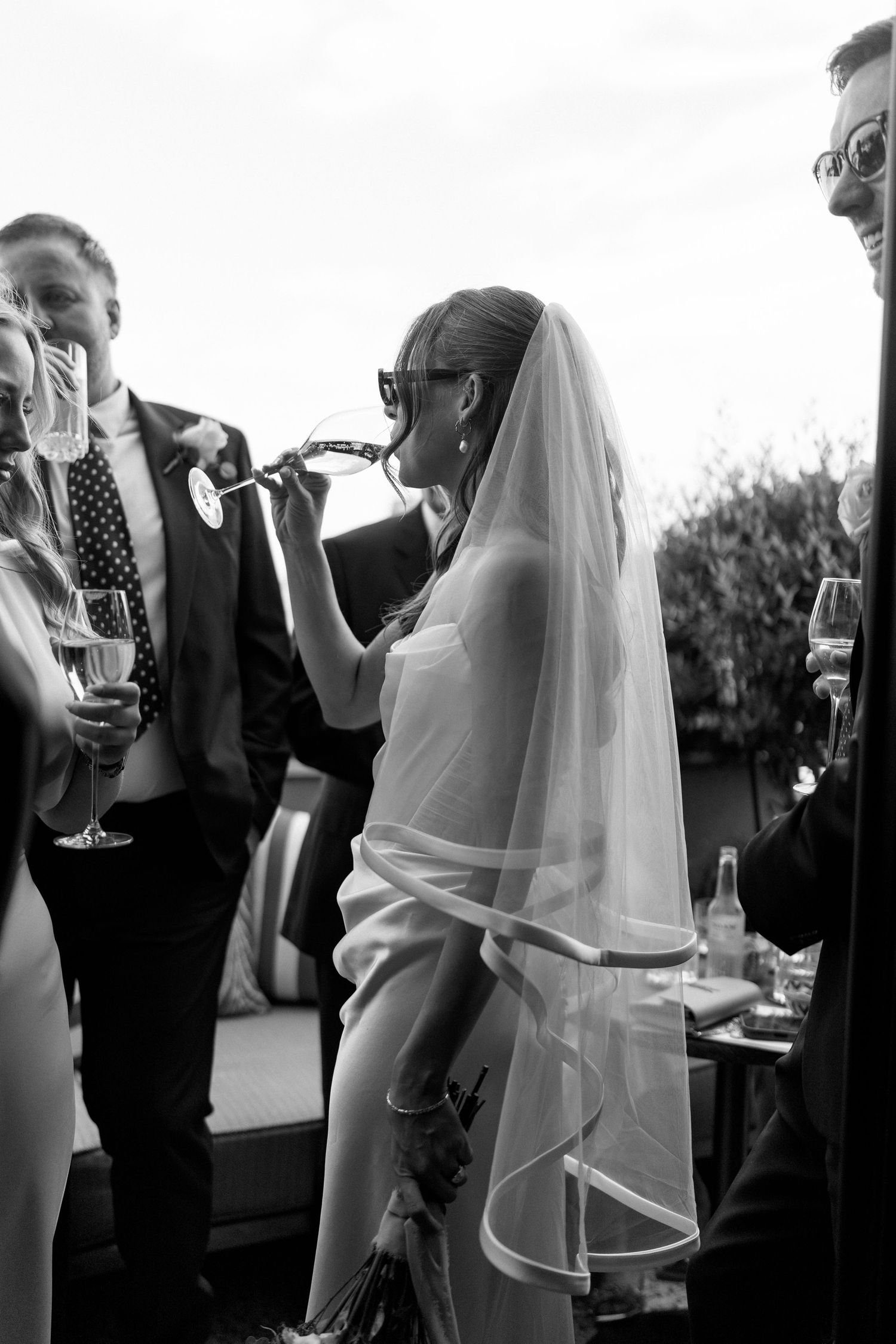 Bride in white gown and veil drinks from bottle at outdoor wedding reception in black and white photo.