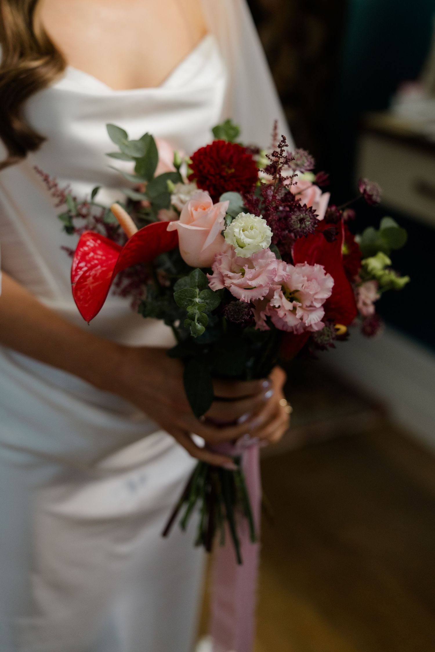Bride holding a lush wedding bouquet with red anthuriums, pink roses, dark dahlias, and greenery in a white gown.