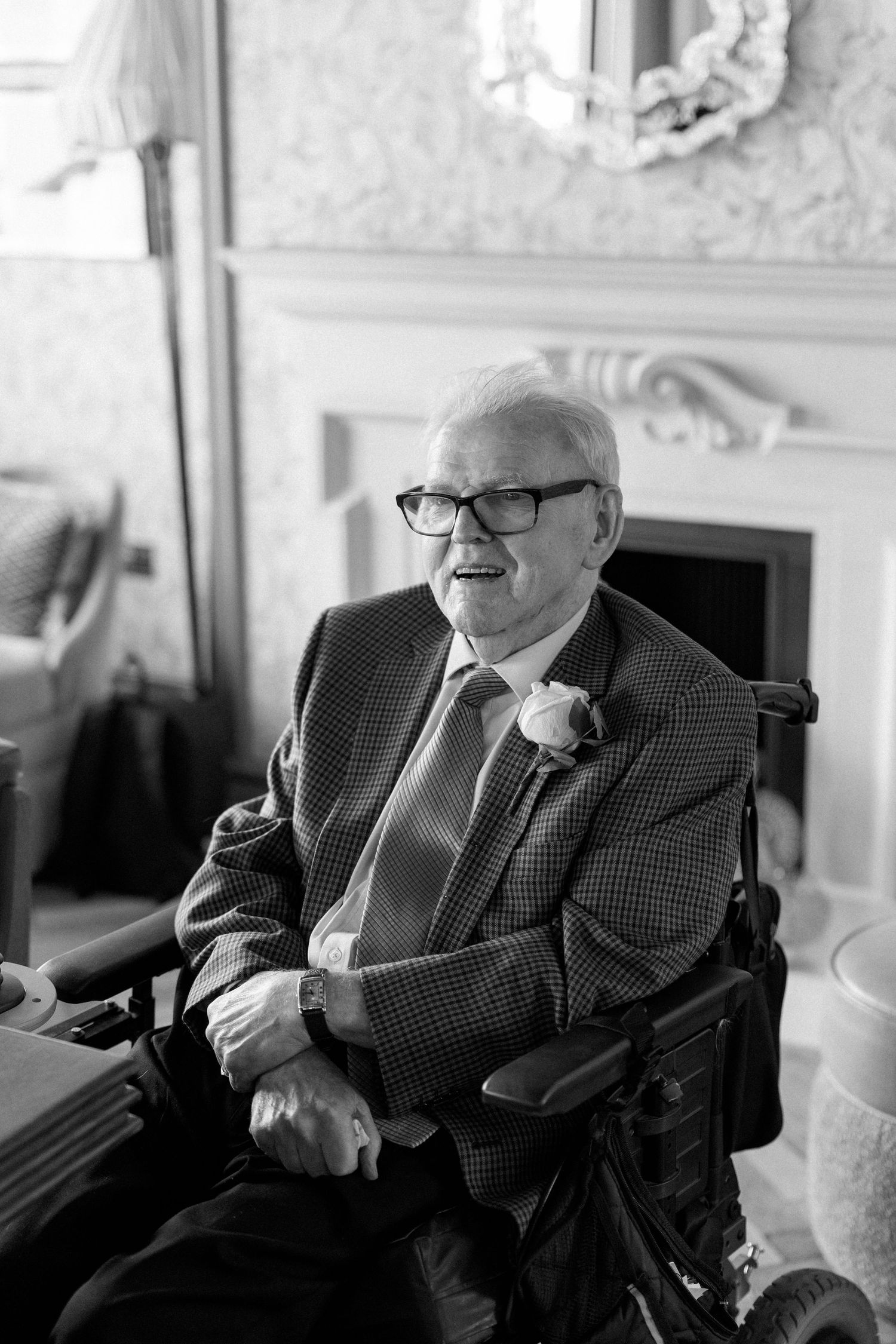 Elderly man in suit with boutonniere smiles warmly while seated in wheelchair in elegant room.