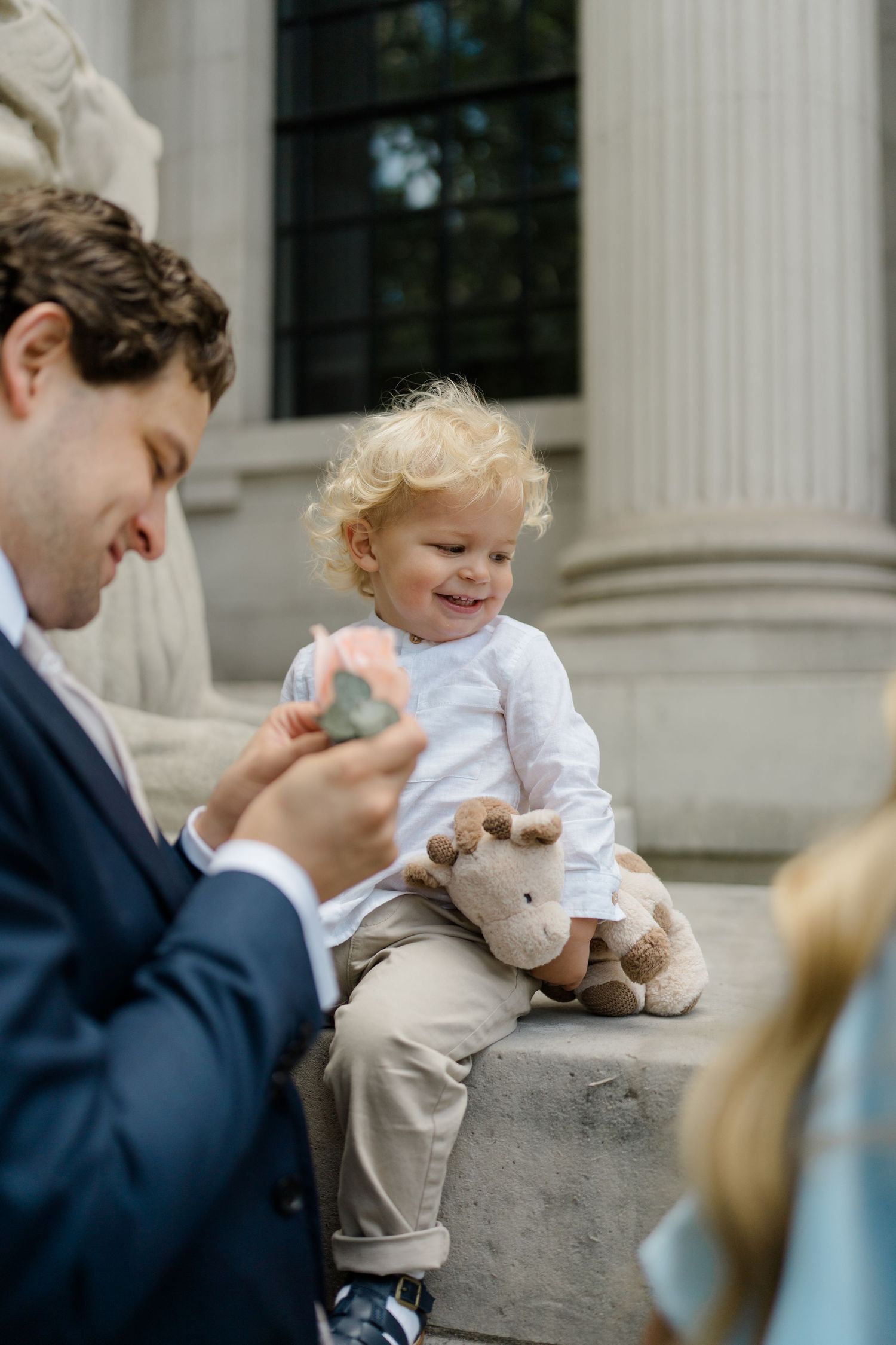 Smiling toddler with curly hair holds stuffed giraffe toy while sitting on stone steps with father.