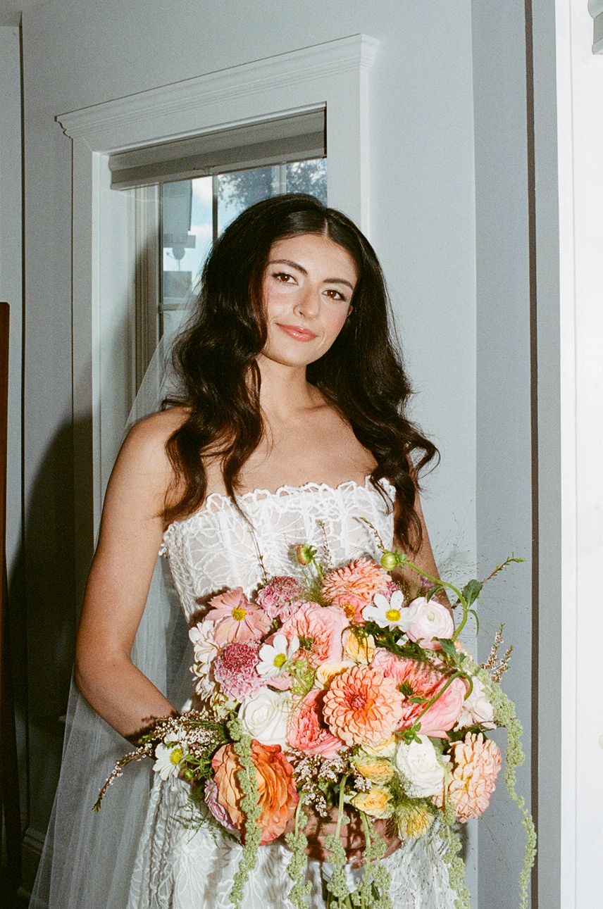 Bride in lace gown holding lush pink and orange floral bouquet with dahlias, standing in soft natural light.