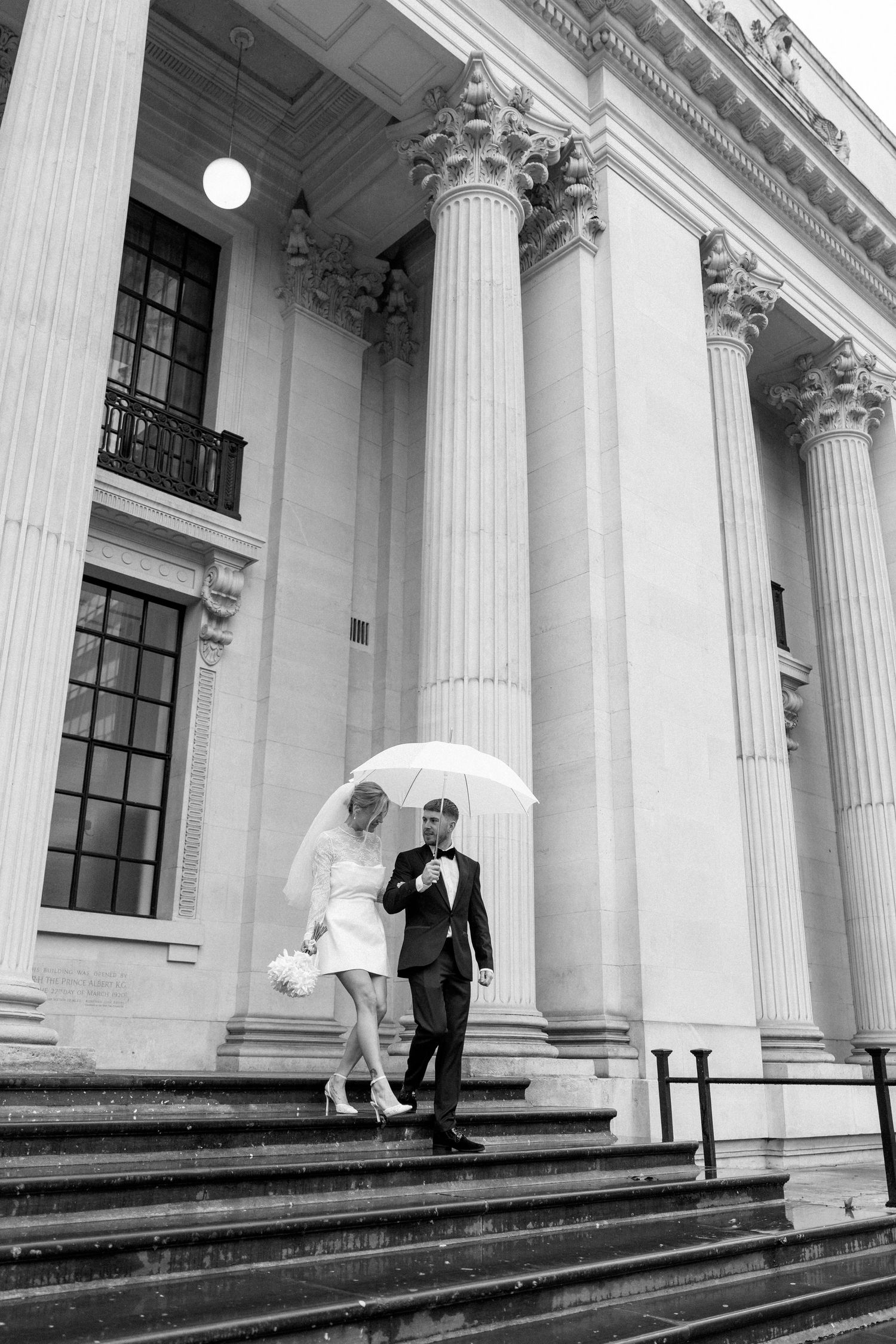 A bride and groom with umbrellas descend grand stone steps with classical columns.