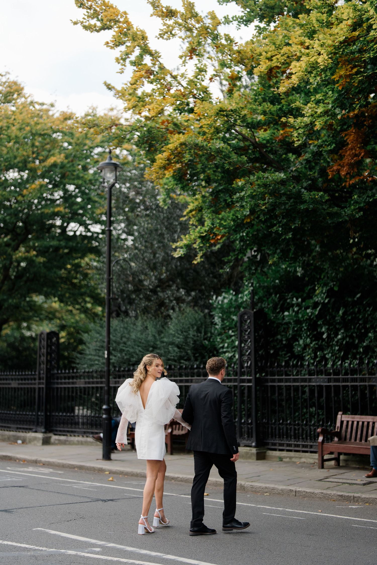 A couple walks along a park path with autumn trees and iron fencing in the background.