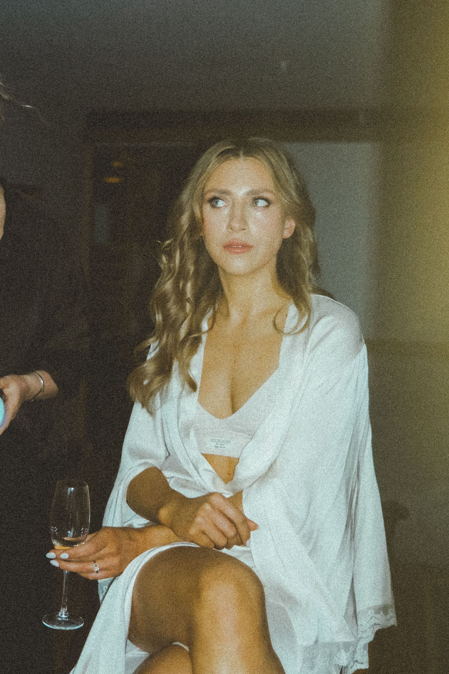 A person in a white silk robe sits elegantly holding a wine glass in a dimly lit room.