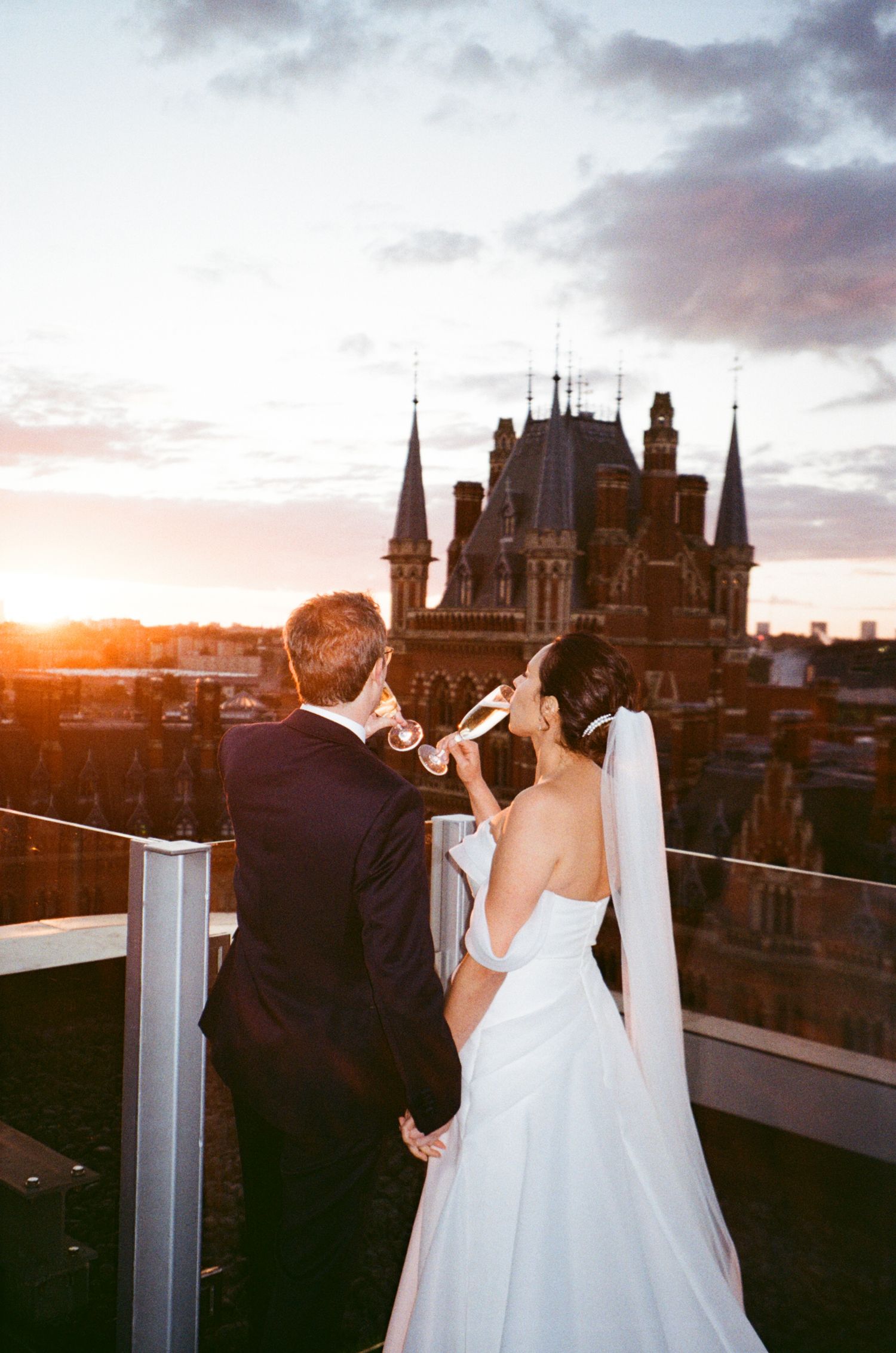 Newlyweds share an intimate moment on a rooftop with gothic architecture silhouetted against sunset sky.