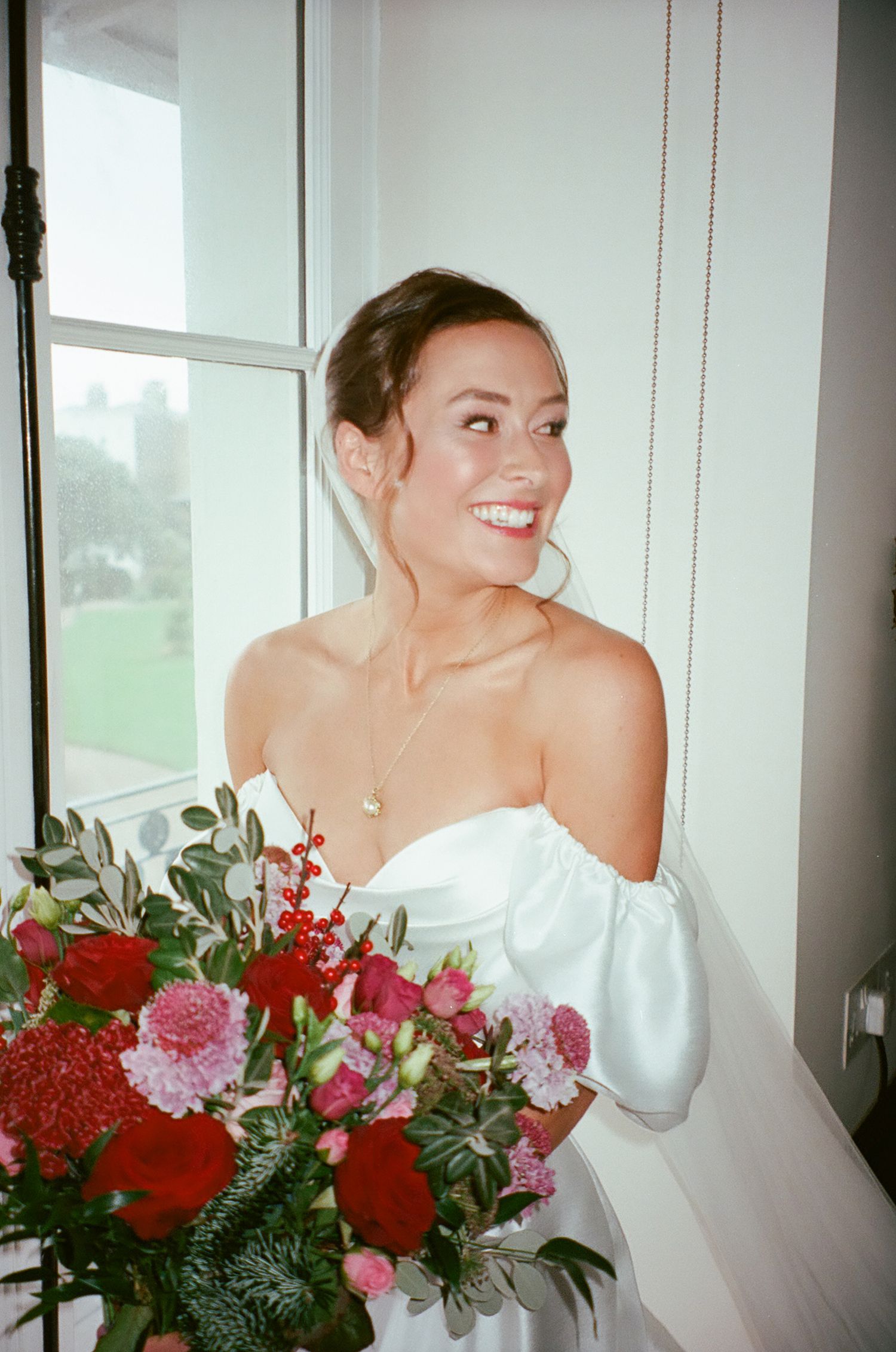 A person in a white off-shoulder gown smiles while holding a vibrant bouquet of red and pink flowers by a window.