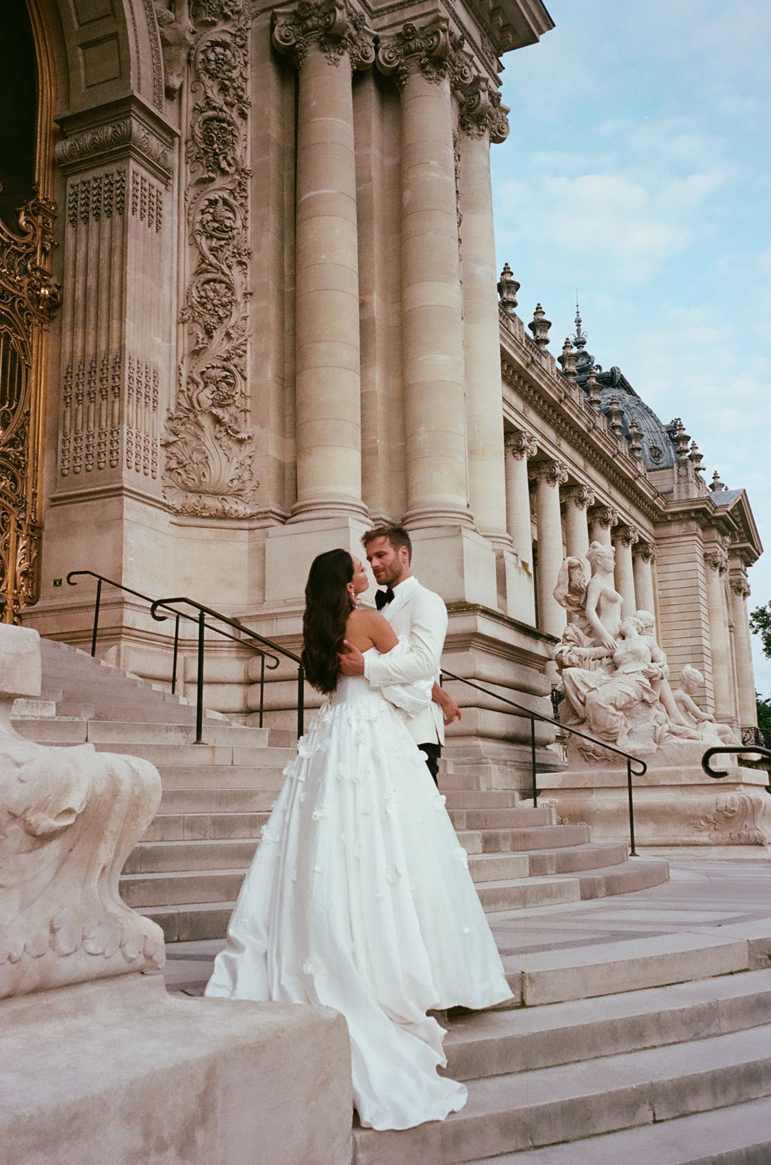 A couple in wedding attire embraces on the grand stone steps of an ornate Beaux-Arts style building.