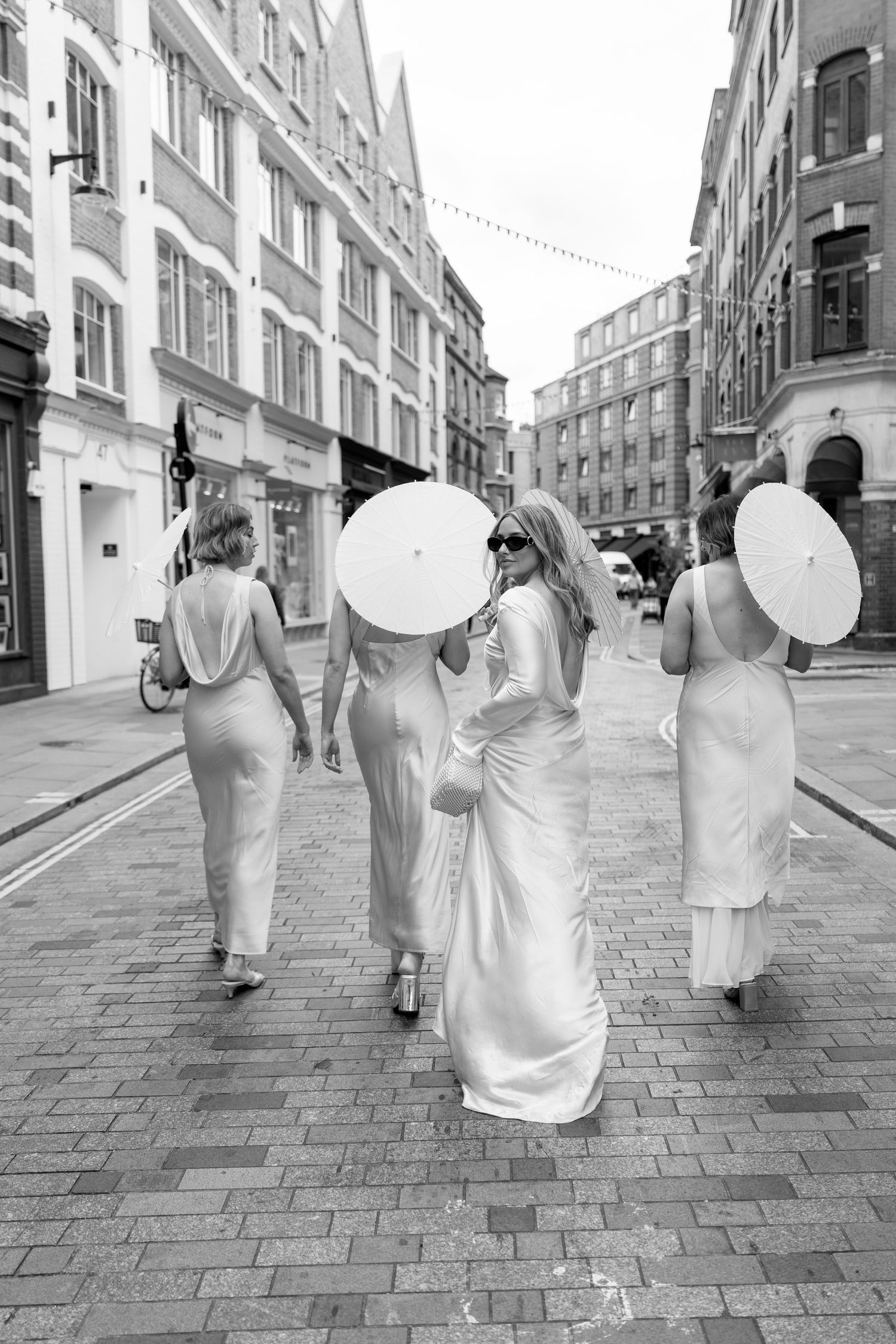 People in white dresses and sun hats walk along a cobblestone street in an artistic sequence.
