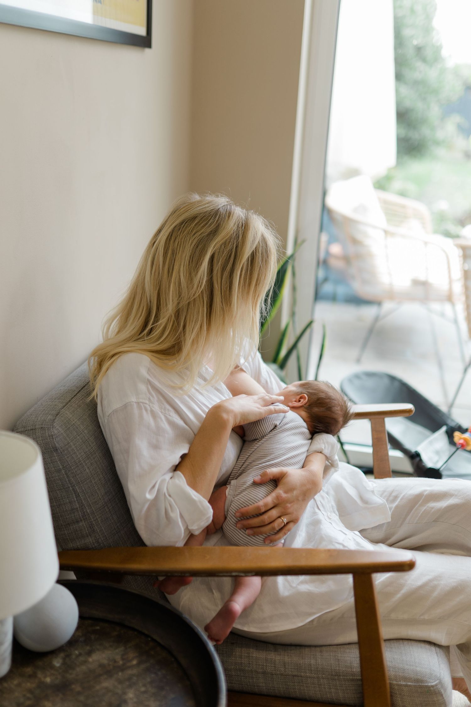 A parent in a white shirt sits in a cozy armchair by a window, cradling and feeding a newborn baby.