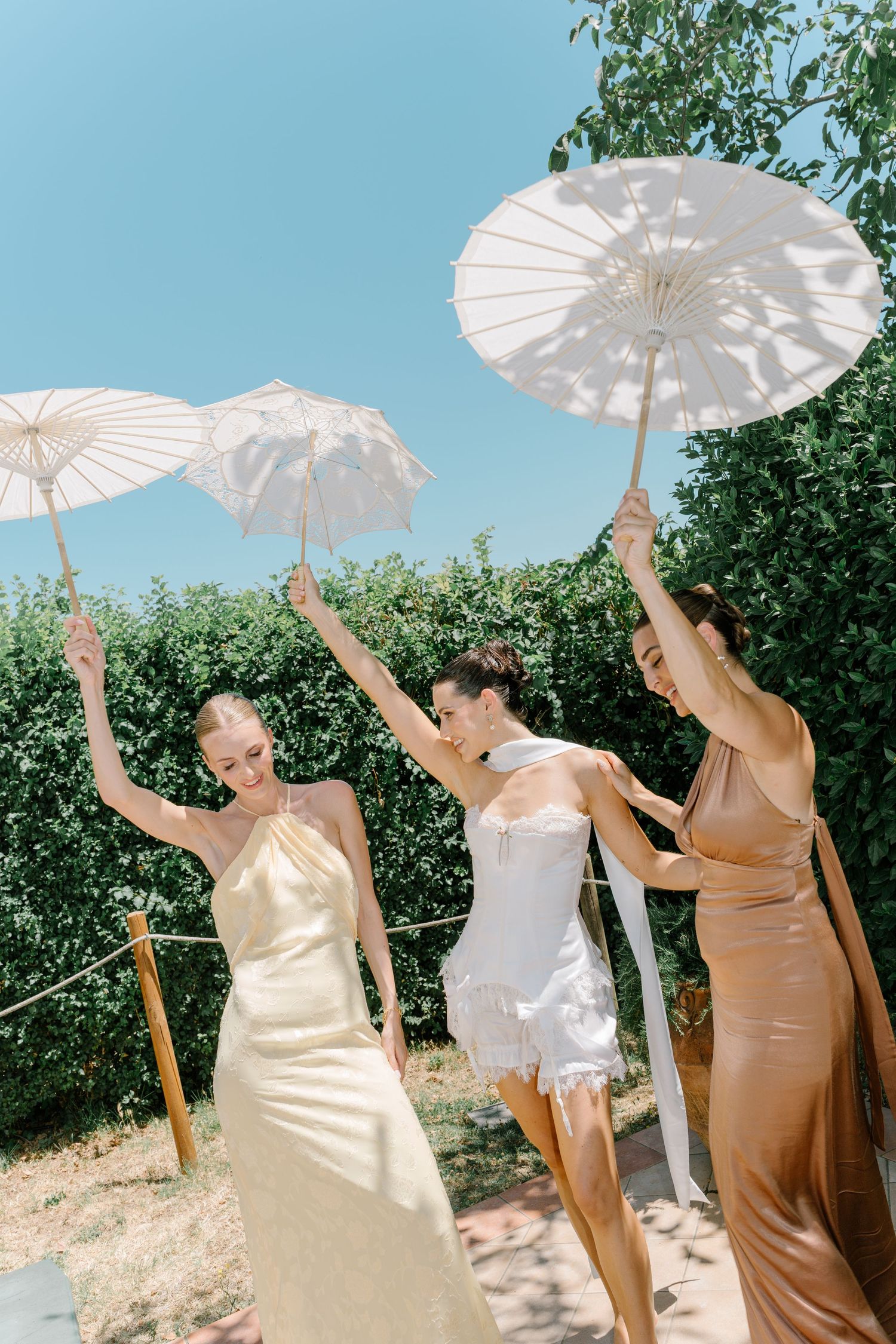 Three people in elegant dresses hold white paper parasols while posing outdoors on a sunny day against green foliage.