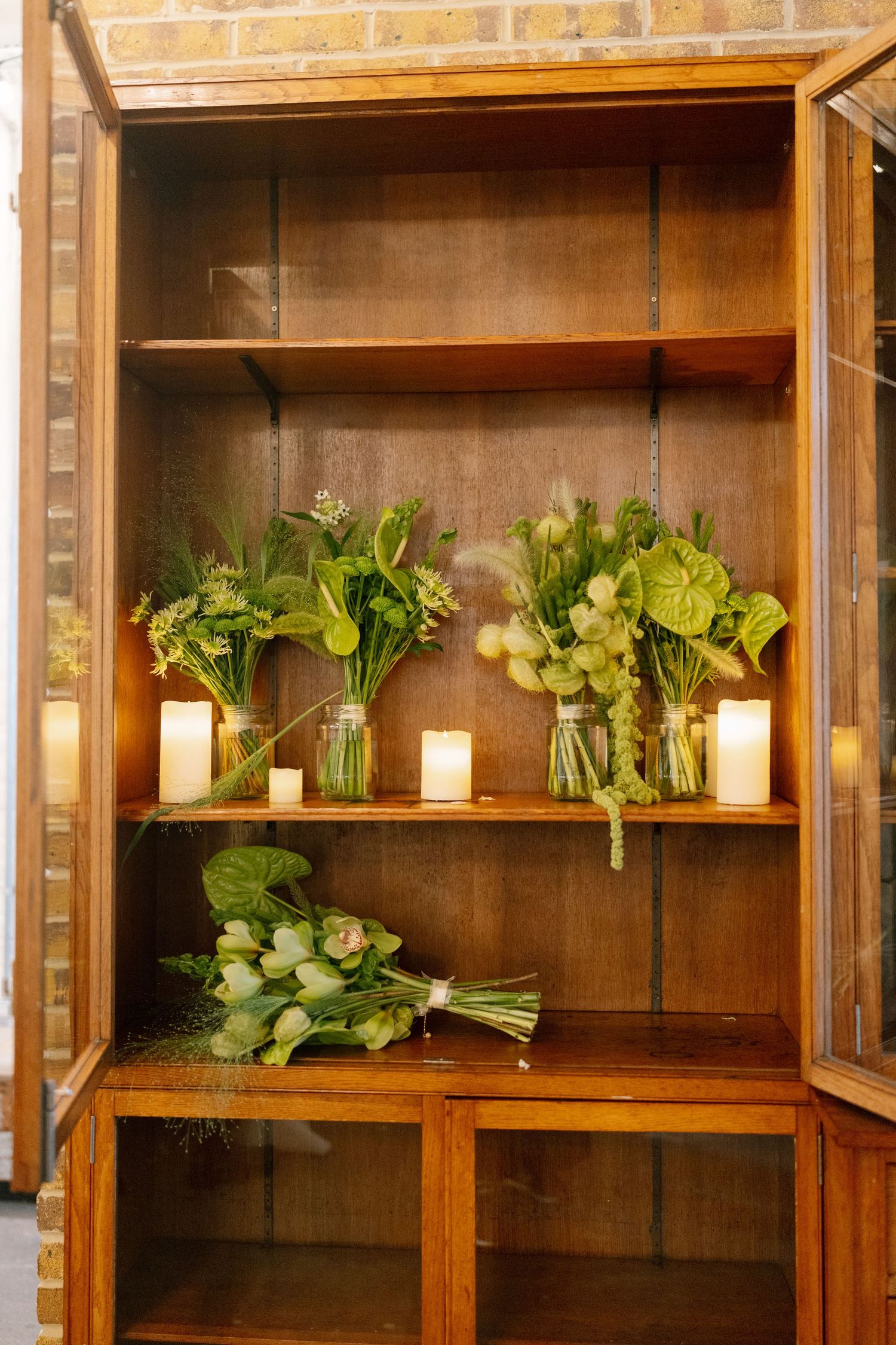 Wooden display cabinet with green plants and candles arranged on shelves.
