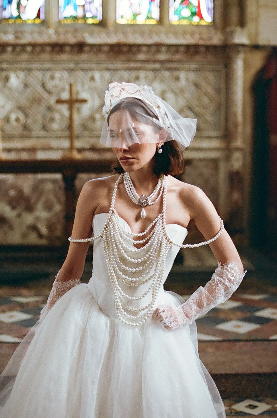 Elegant bride in white tulle gown with draped pearl necklaces and lace gloves poses in ornate church interior.